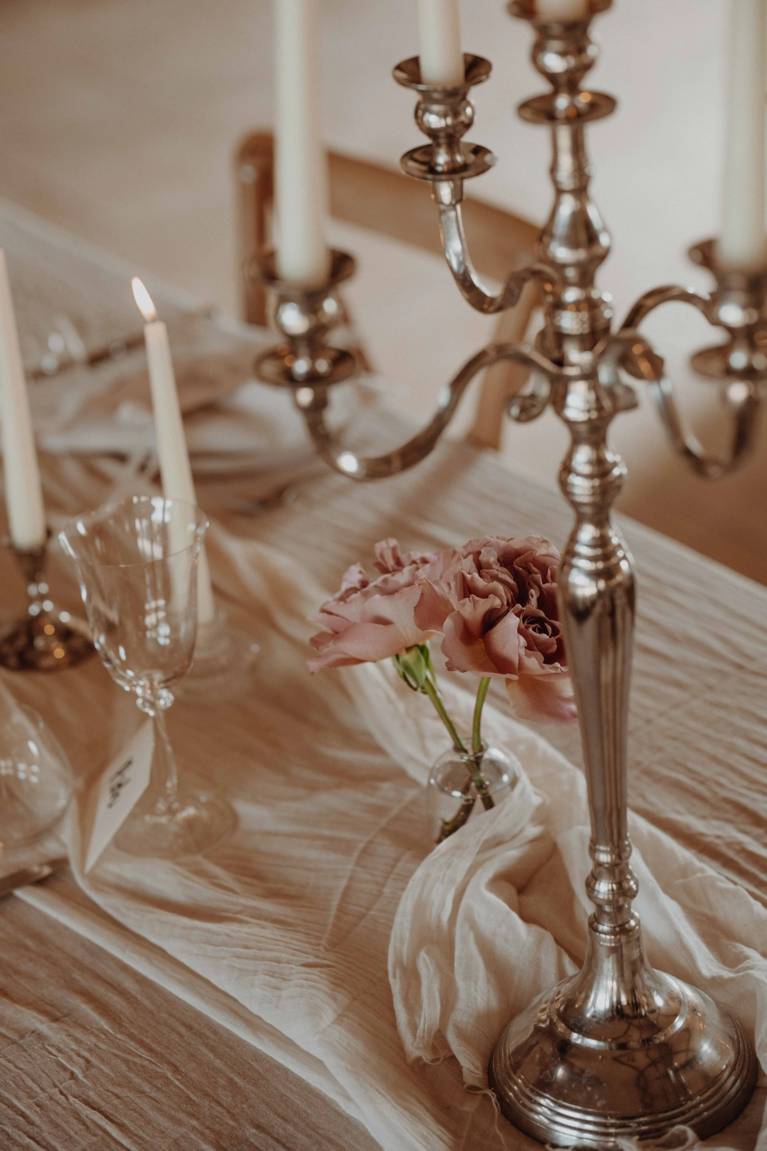 A table decorated with a silver candelabra holding lit candles, a glass vase with pink flowers, a folded white cloth, and empty wine glasses.