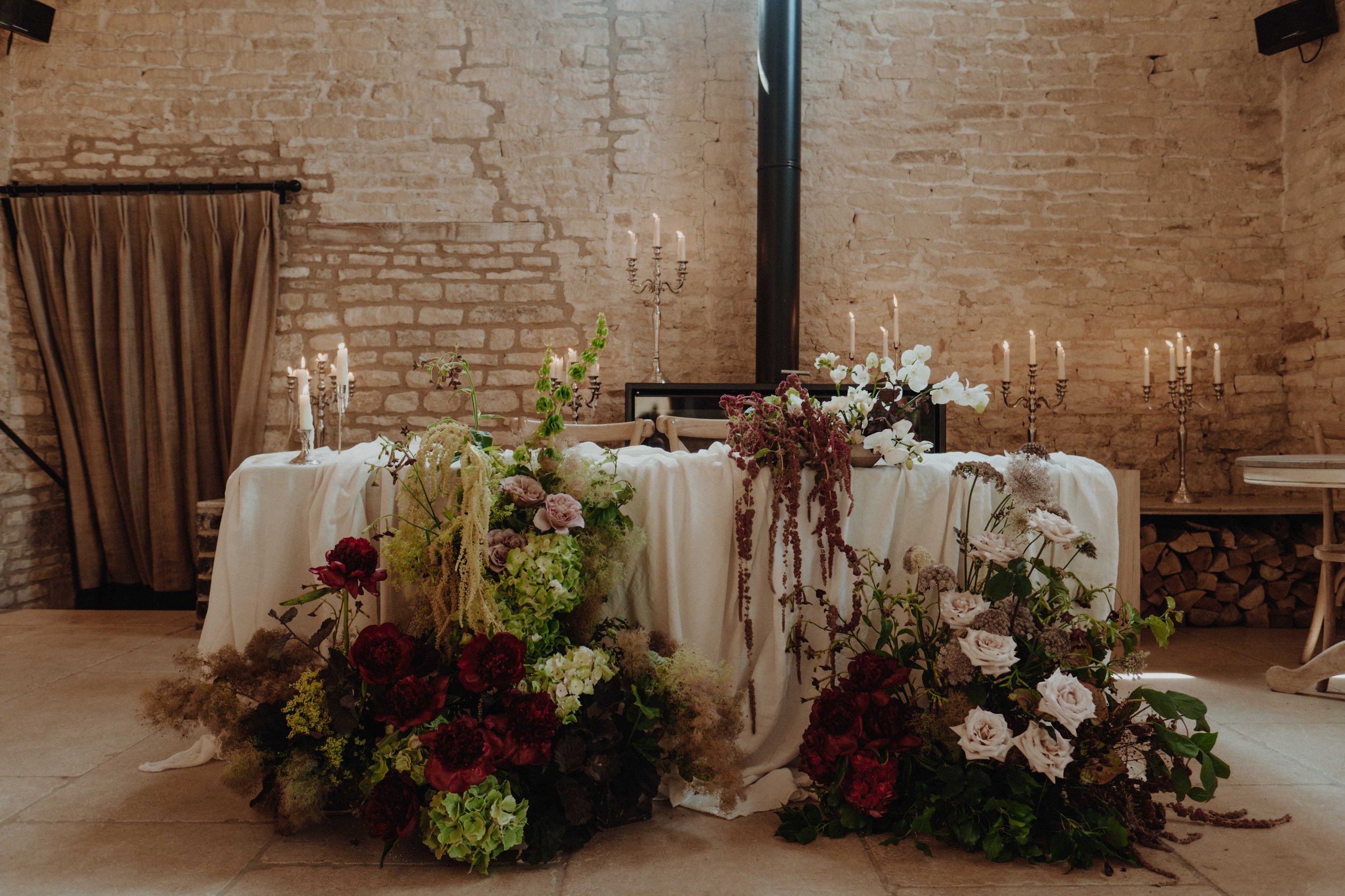 Elegant wedding reception setup with a long table covered in a white cloth, decorated with multiple floral arrangements including roses, peonies, and greenery, and illuminated by candlelight in silver candelabras. The setting is indoors with exposed 