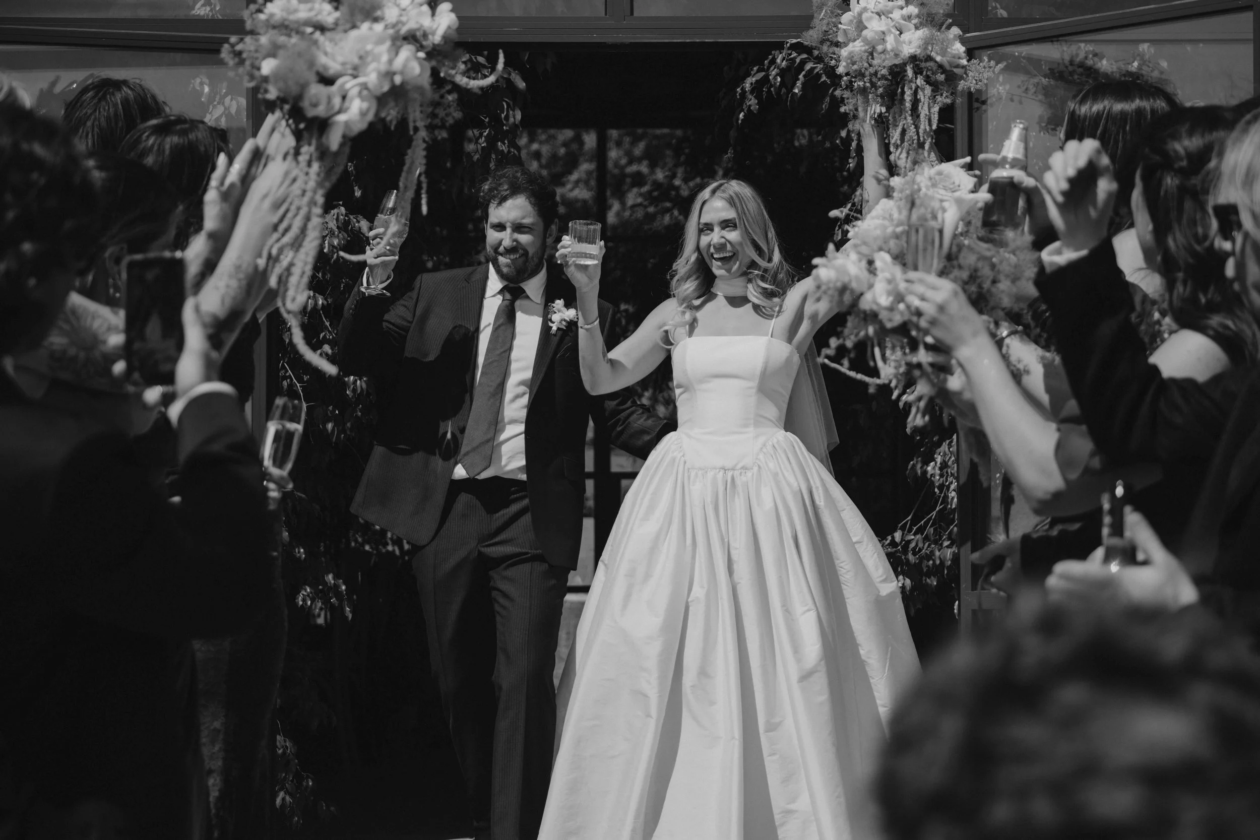A wedding celebration with a bride and groom surrounded by friends, holding glasses, and smiling as they walk through an arch decorated with flowers.