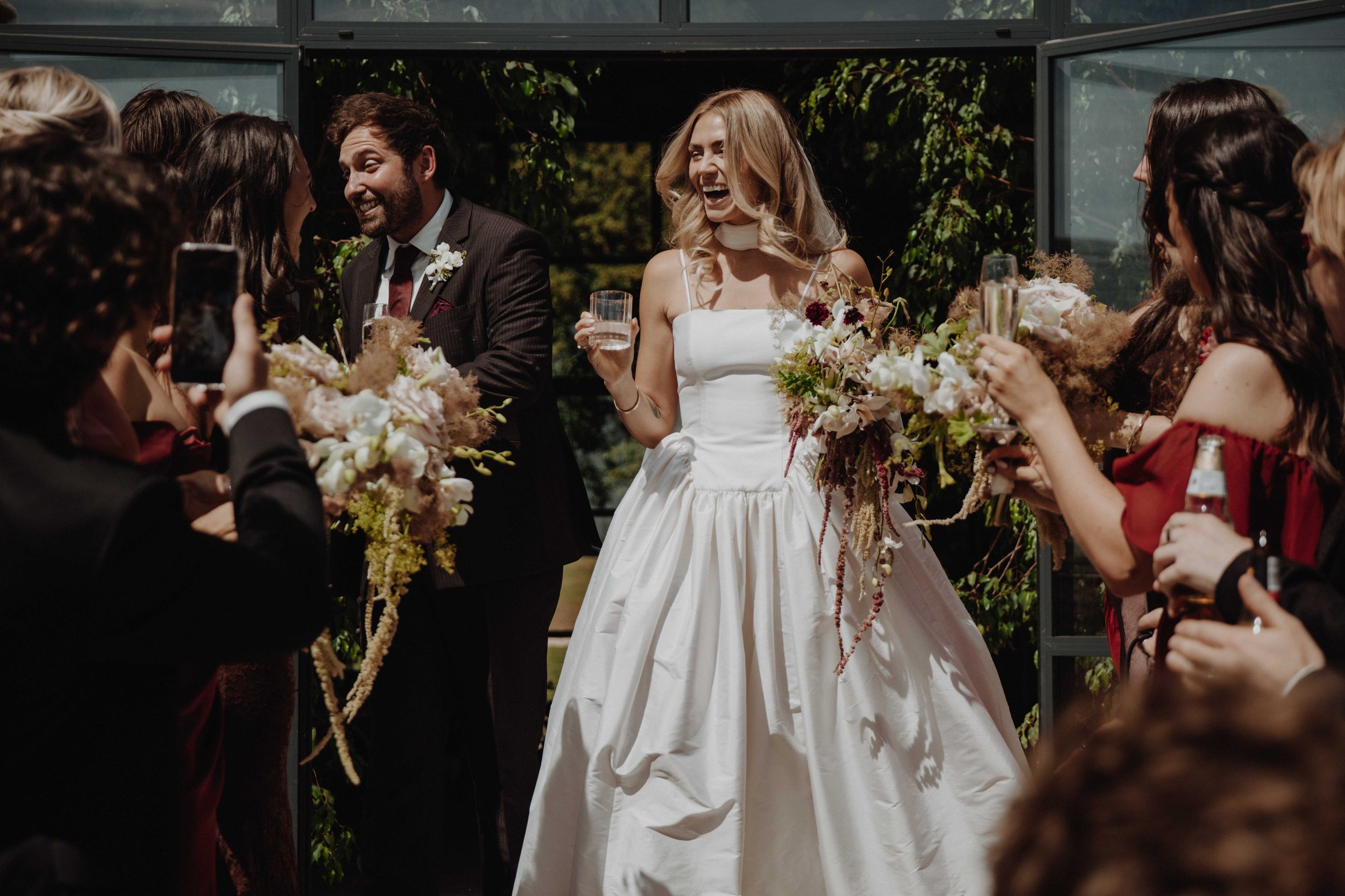 Wedding celebration with bride holding a glass, surrounded by friends and family, outdoors.