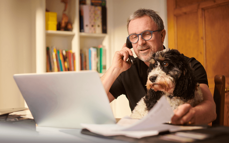Business owner working at a laptop with paperwork and a dog beside him, representing business support and bookkeeping help in New Zealand