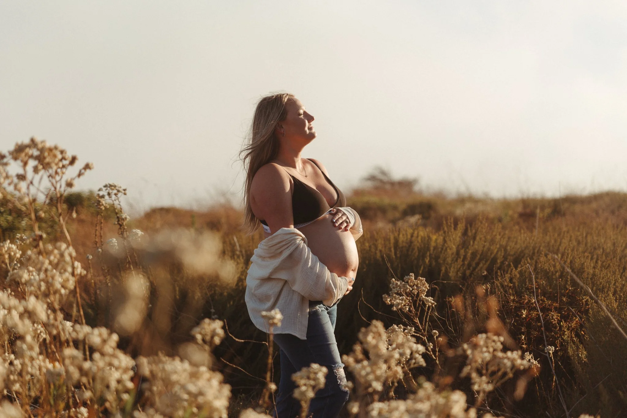 Pregnant person standing in a field of flowers, wearing a sports bra and jeans