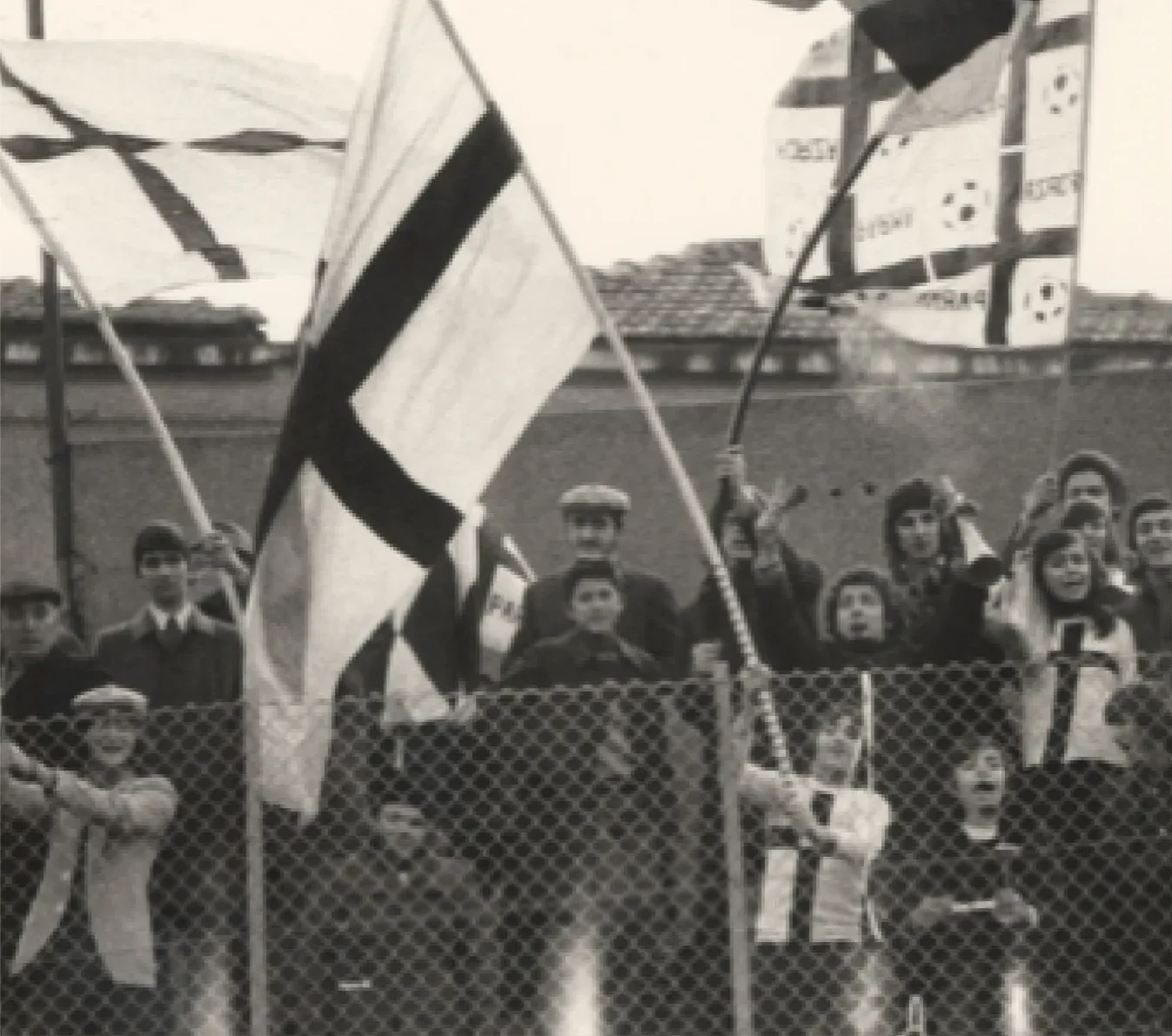 A group of people standing behind a chain-link fence, holding flags, with some children in the front. The scene appears to be a protest or gathering, in black and white.