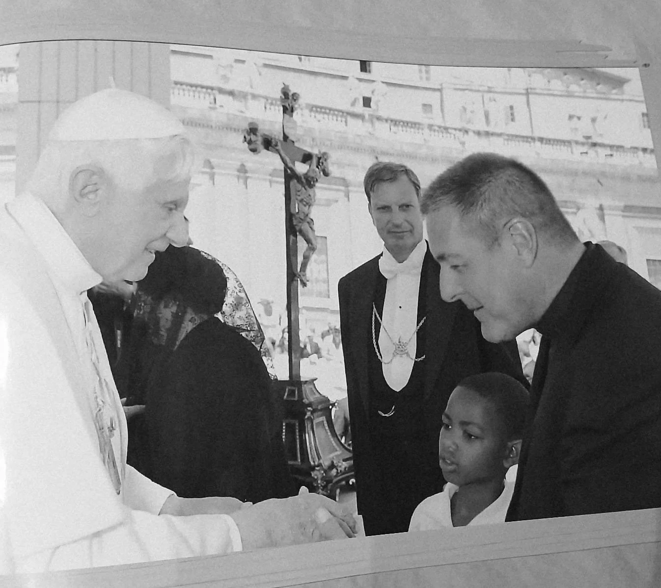 Black and white photo of Pope Francis shaking hands with a young boy, with two men in tuxedos and a woman in a black dress nearby, in an outdoor setting with a large historic building in the background.