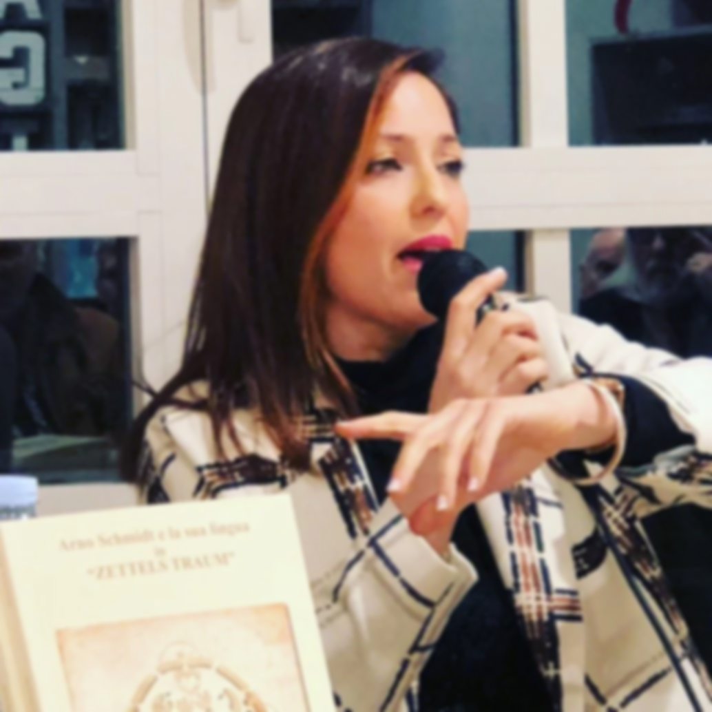 A woman with brown hair is singing into a black microphone indoors, with her eyes partially closed and a focused expression. She is wearing a beige blazer with a black and white pattern. In front of her, there is a photo or book with the title in Spanish visible.
