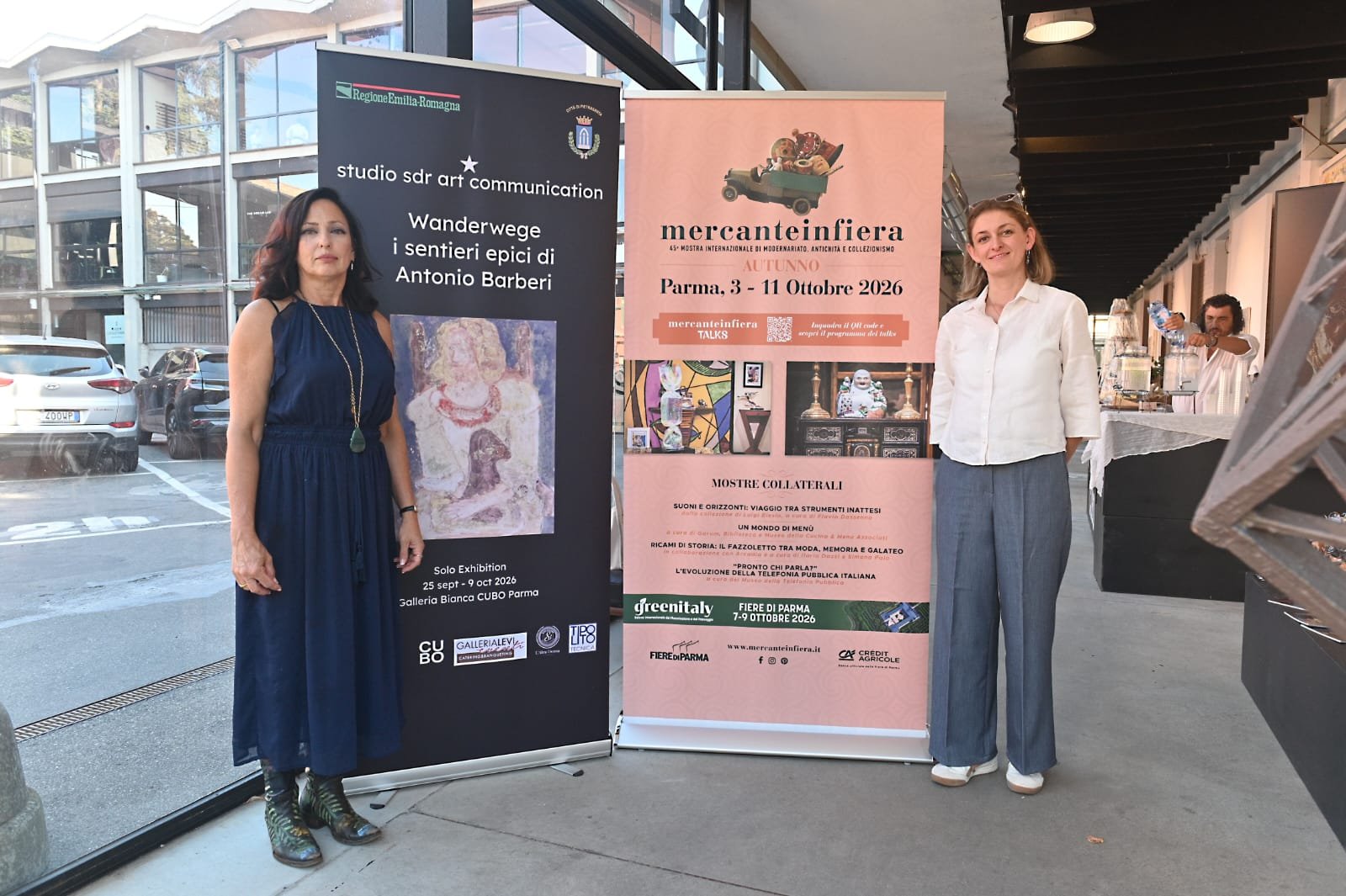 Two women standing next to each other in front of pink and black exhibition banners, inside a building with glass walls and parked cars outside.