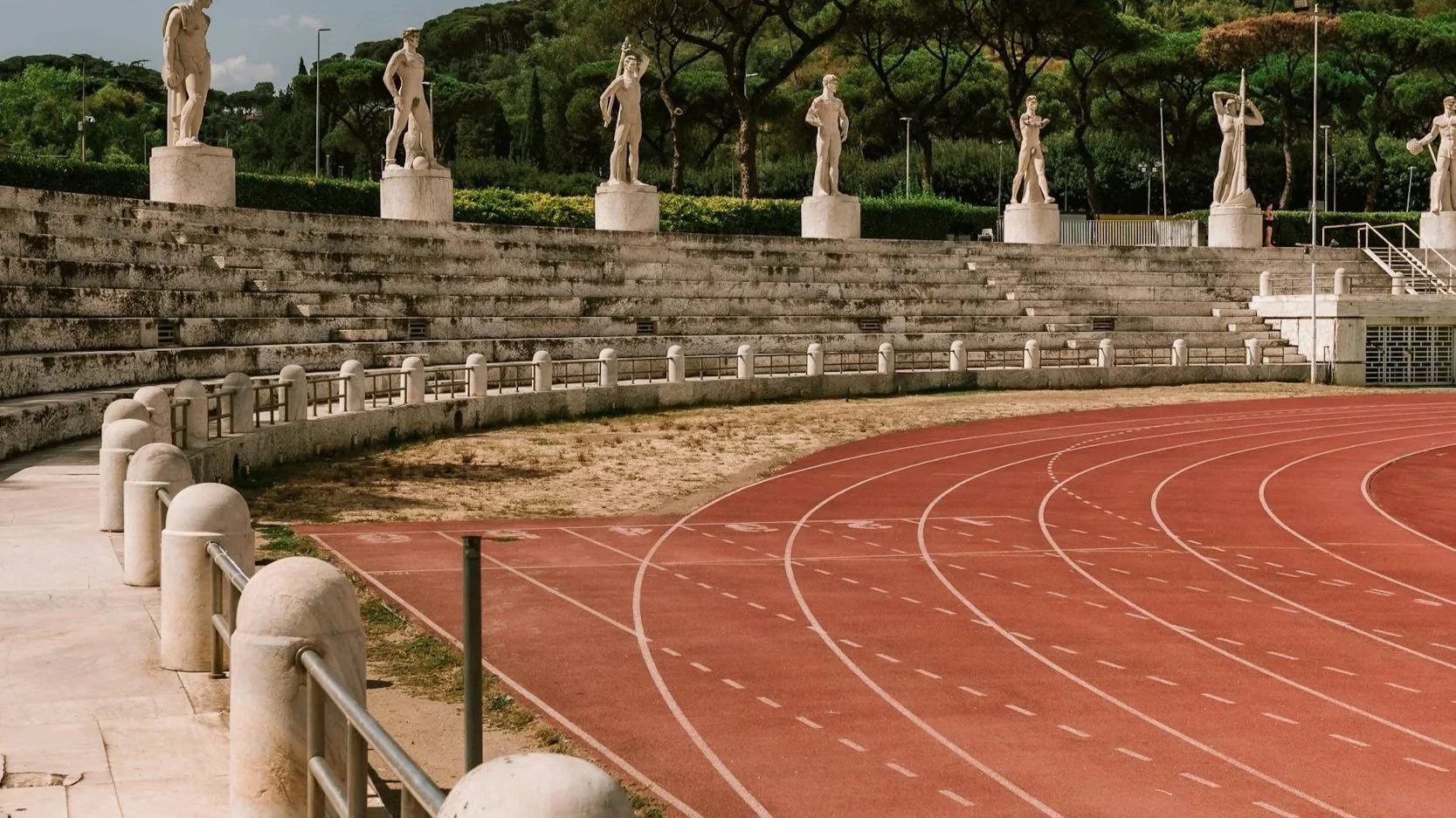 An empty outdoor running track with a curved red surface and white lines, surrounded by stone bleachers and statues of classical figures, with trees and a hill in the background.