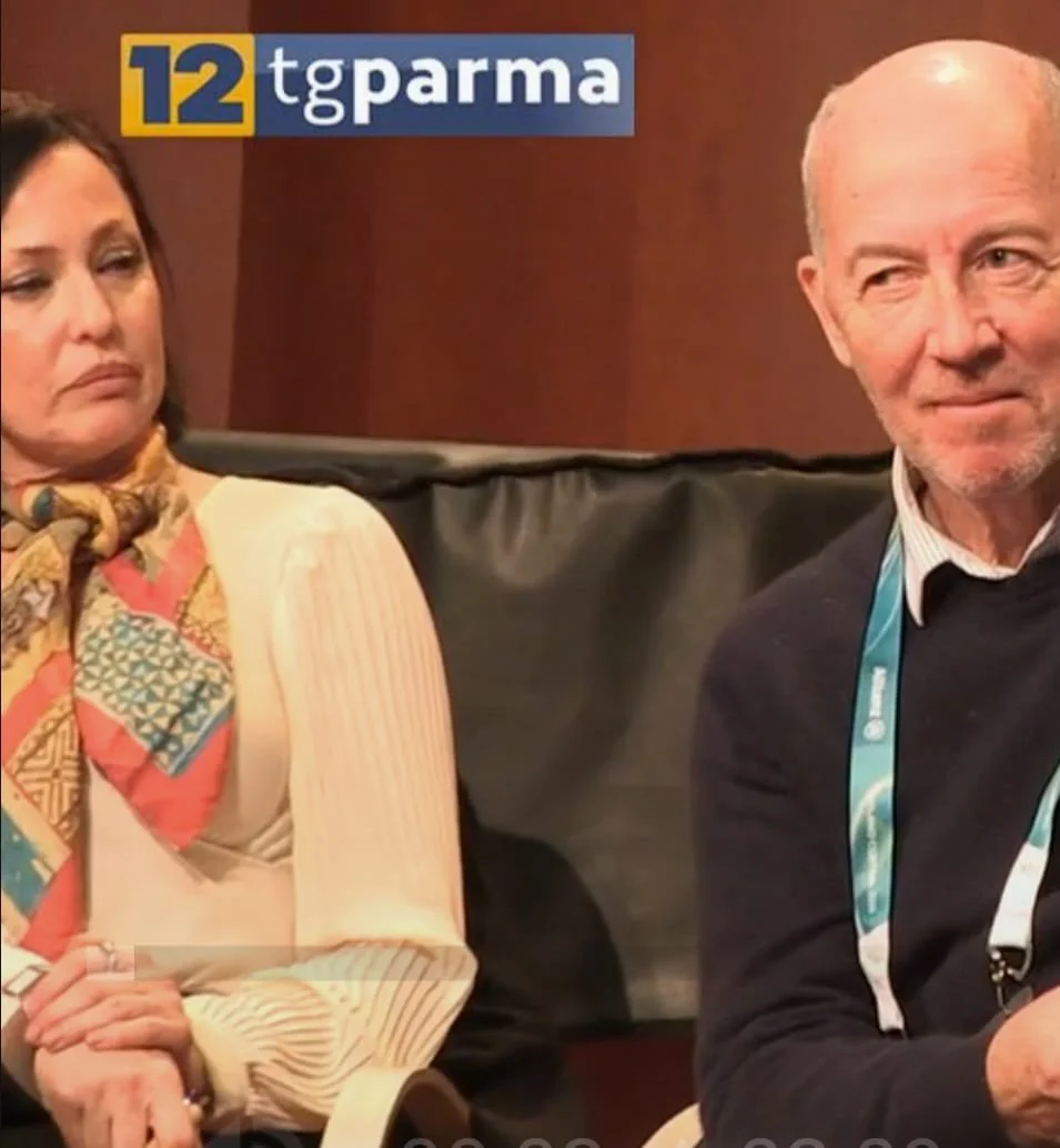 Two people sitting on a couch during an interview on 'Tg Parma' news segment. The woman on the left wears a cream-colored blouse and a colorful scarf, appears serious. The man on the right wears a dark sweater over a collared shirt, has a sympathetic expression. A banner in the top left displays '12 tg parma'.