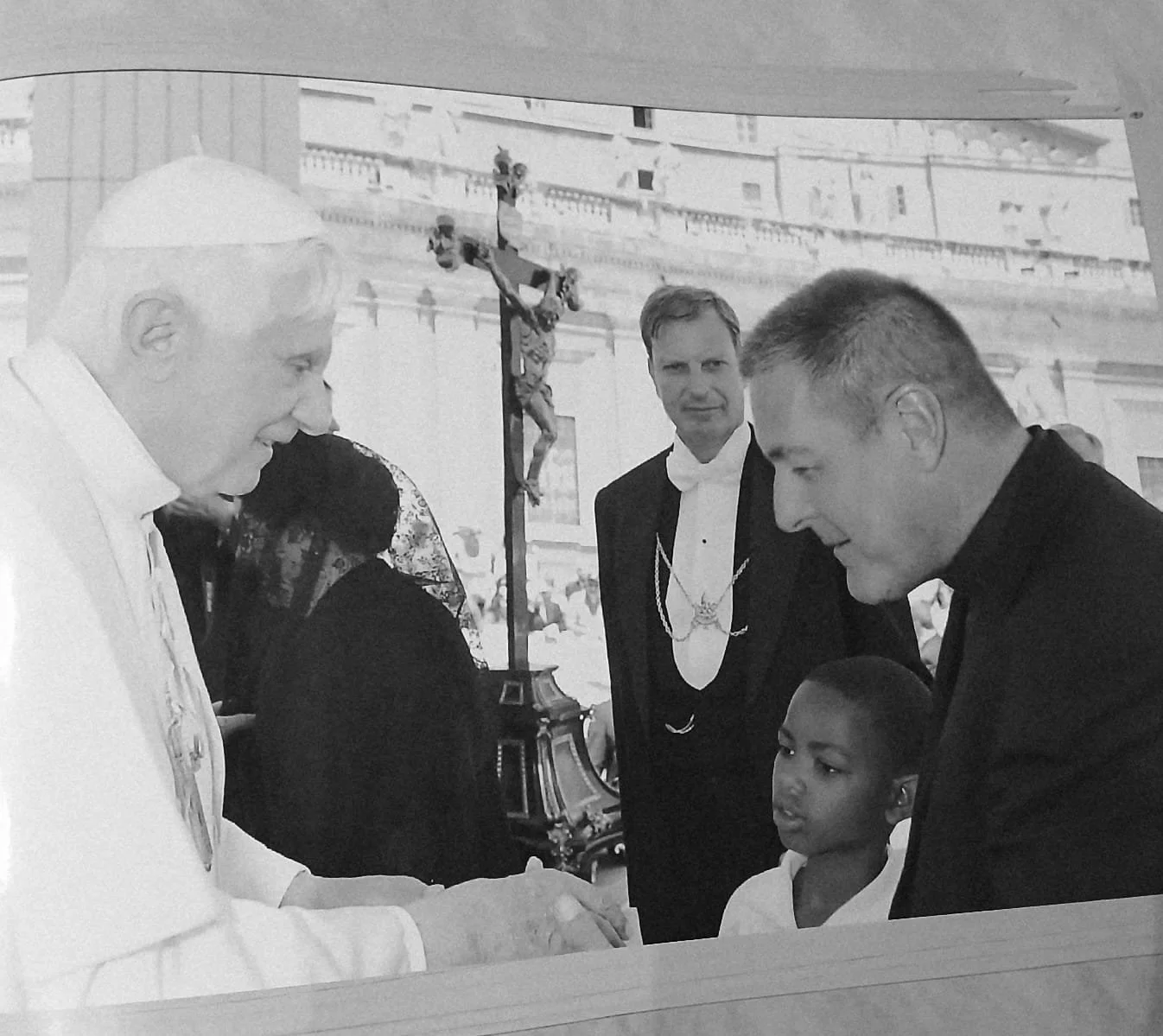 Pope Francis shaking hands with a young boy during an outdoor religious event, with two men in tuxedos and a religious statue of Jesus on the cross visible in the background.