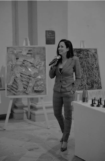 Woman in a suit speaking into a microphone at an art gallery or exhibition.
