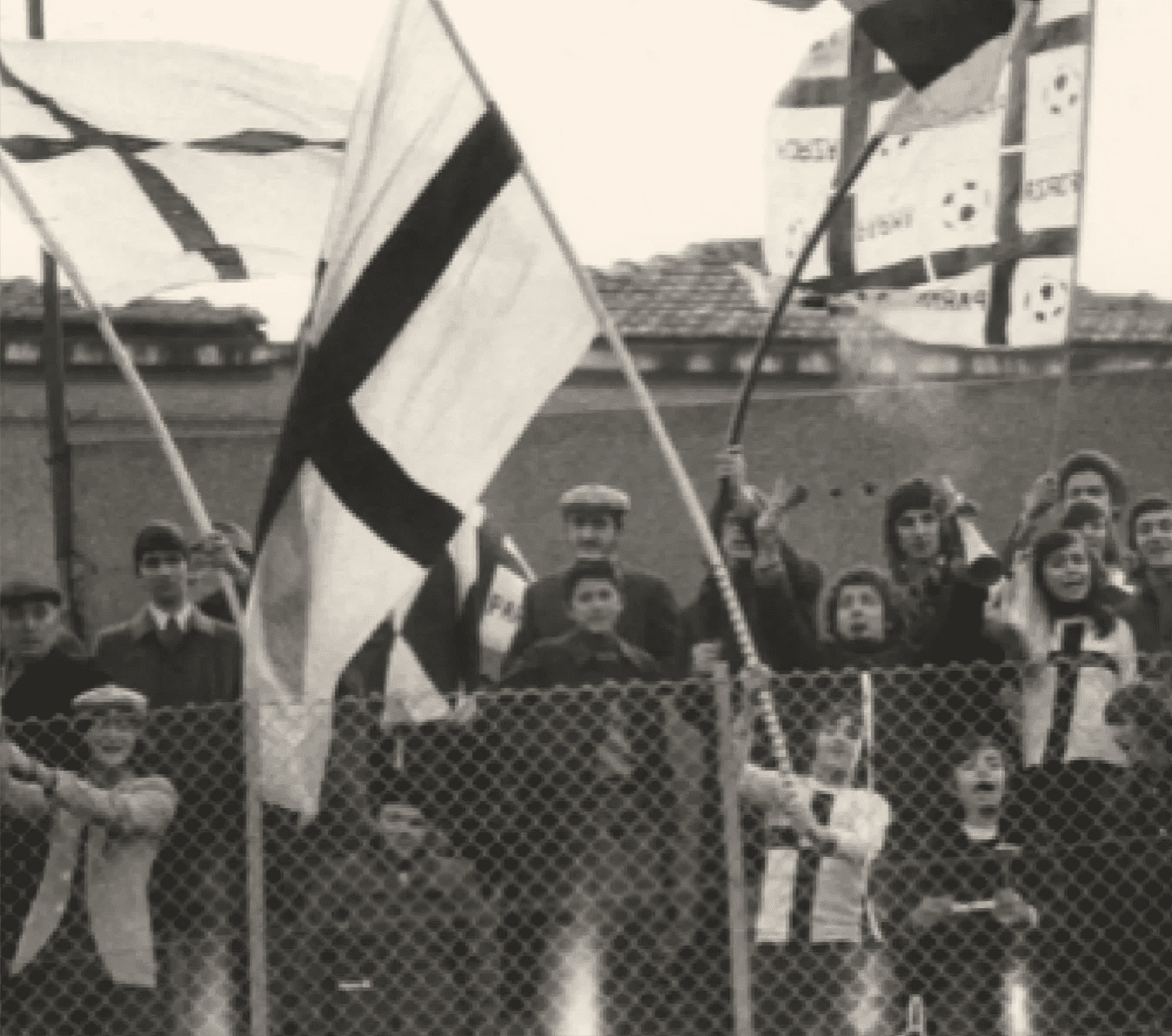 Children and adults holding flags behind a chain link fence, celebrating or participating in an event outdoors.