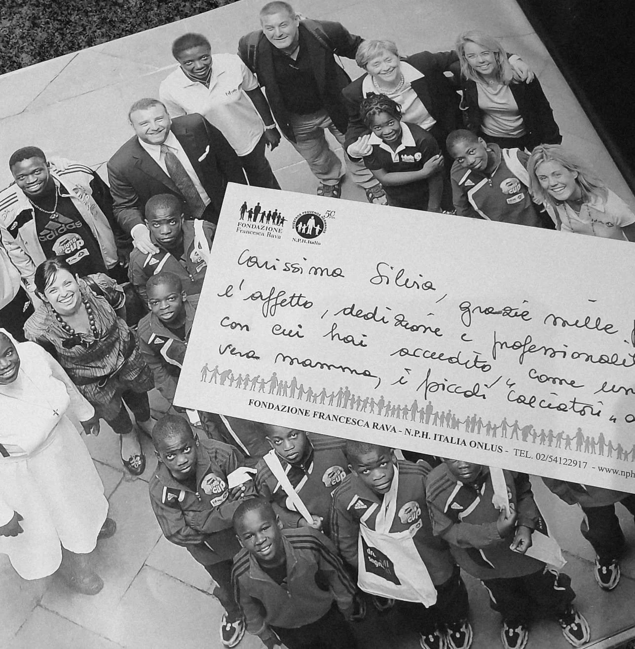 Black and white photo of a diverse group of children and adults standing together, some smiling and looking up, with a handwritten note in Italian on top of the group.