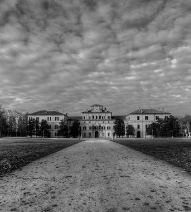 A grand historic mansion with a central entrance, surrounded by trees, set against a cloudy sky in black and white.