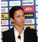 A woman with dark hair in a professional outfit speaking at a press conference with a backdrop of sponsor logos.