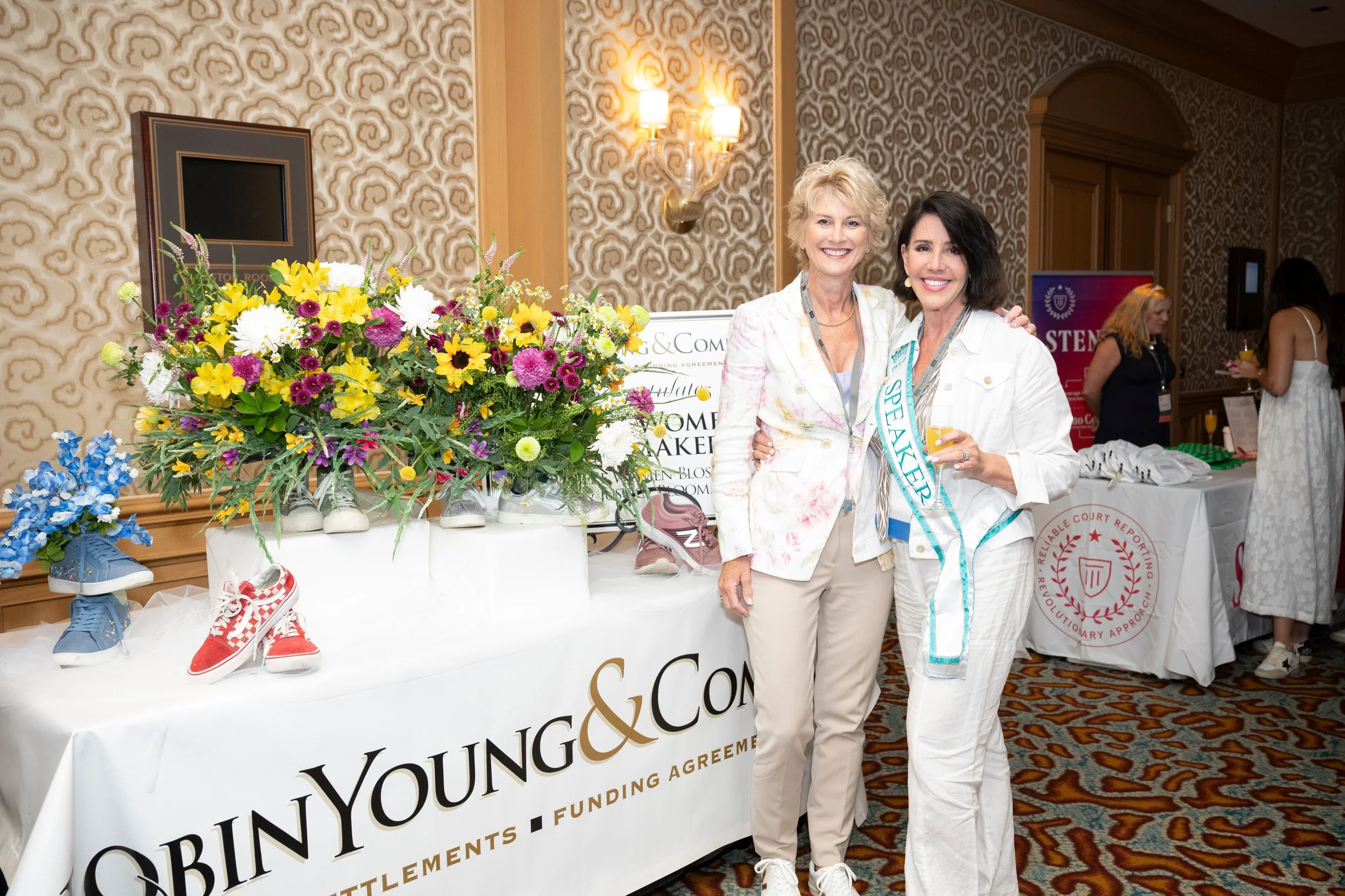 Two women standing together at a conference or event, smiling, with one wearing a speaker sash, standing in front of a table with a large bouquet of colorful flowers and shoes displayed on it.