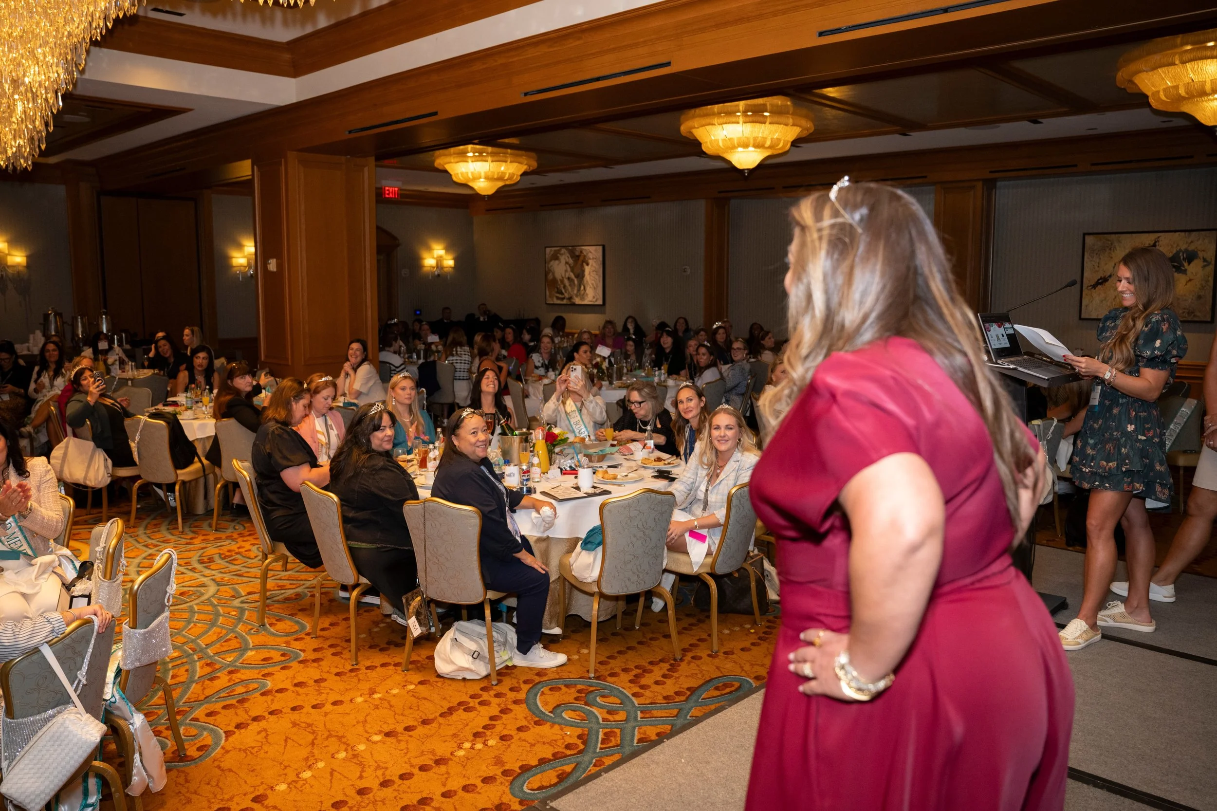 A woman in a red dress stands in front of a large group of women seated at round tables in a banquet hall, with a woman at a keyboard on stage.