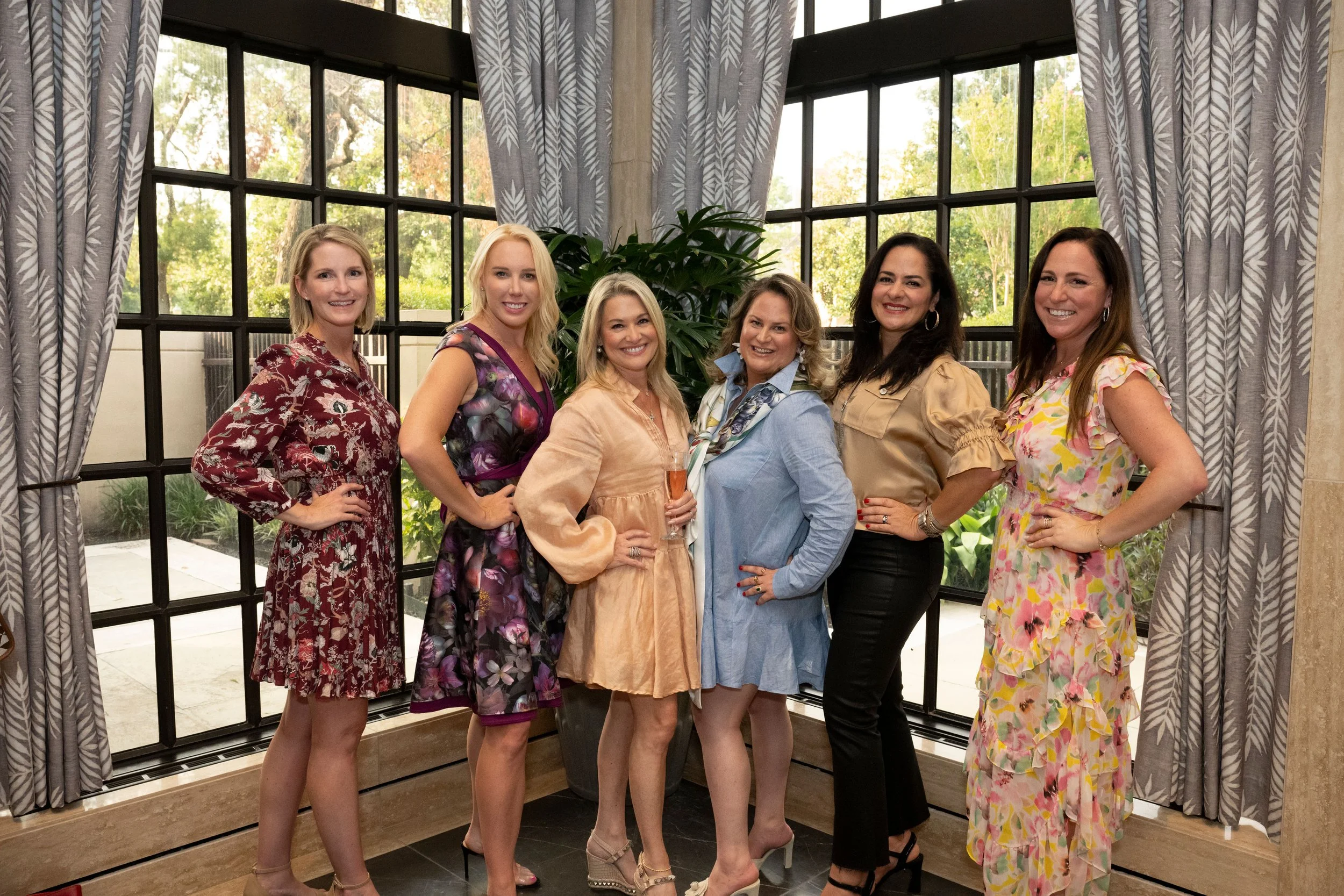 Seven women standing together indoors near large windows with curtains, posing for a photo, with trees visible outside.