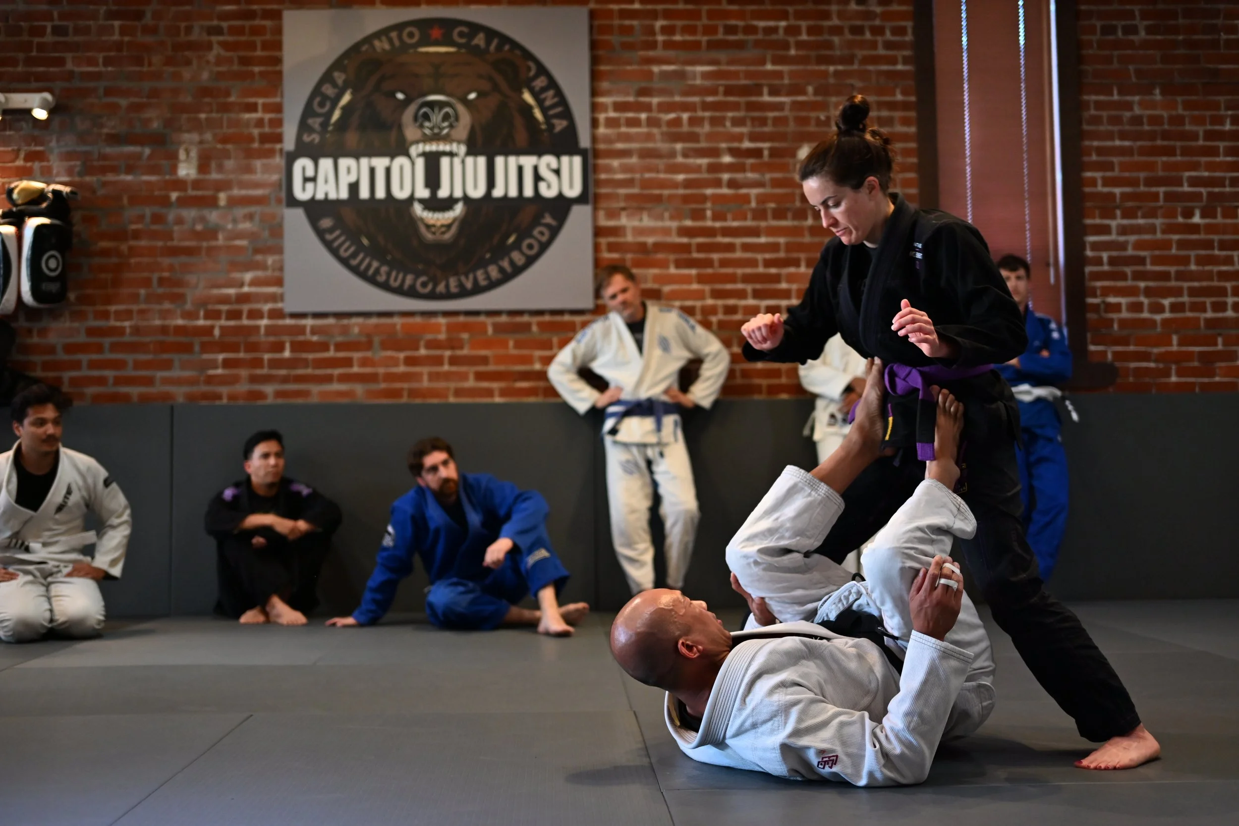 A female martial arts instructor demonstrating a Brazilian Jiu-Jitsu move on a male student in a gym with a group of observers. The instructor is wearing a black gi with a purple belt and is standing over the student, who is lying on his back. The gym has a brick wall with a large Capitol Jiu Jitsu logo. Several students in gis are watching while sitting or standing.
