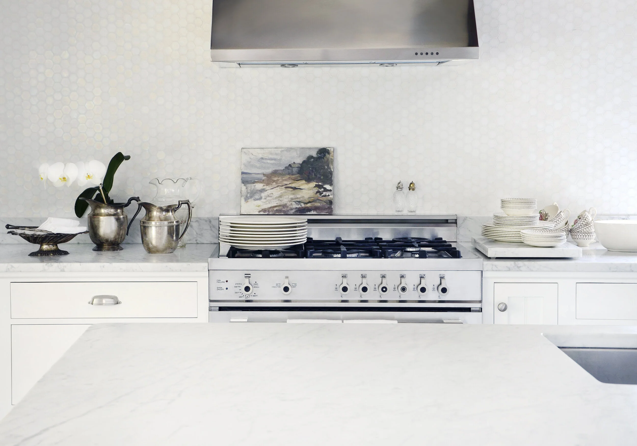 A modern kitchen with a white marble countertop, stainless steel appliances, and a white hexagonal tile backsplash. There are stacked plates, cups, and decorative items on the counter, and a small painting is mounted on the wall above the stove.