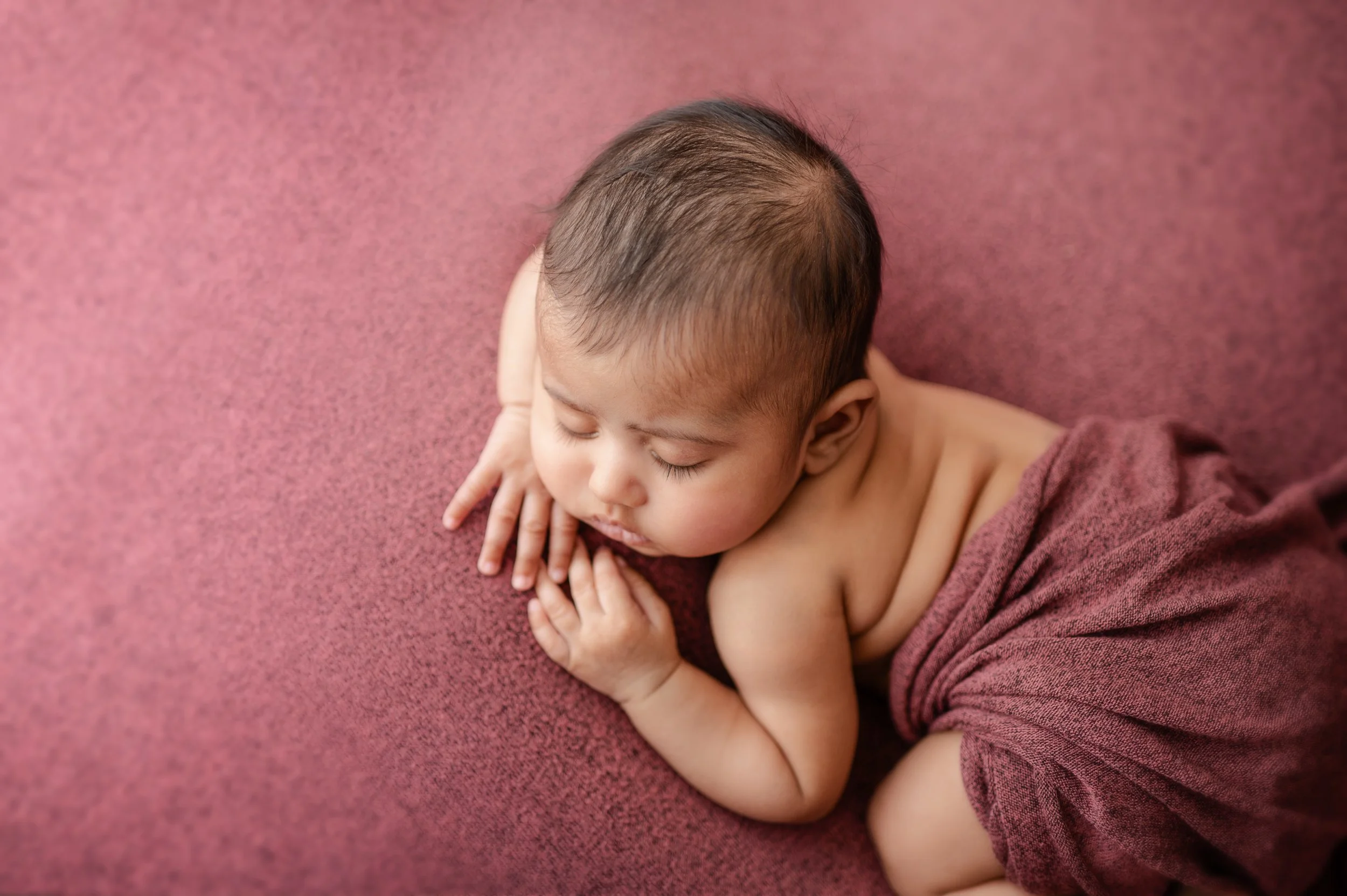 Baby girl wrapped in pink fabric, laying on matching pink fabric asleep with face resting on arms