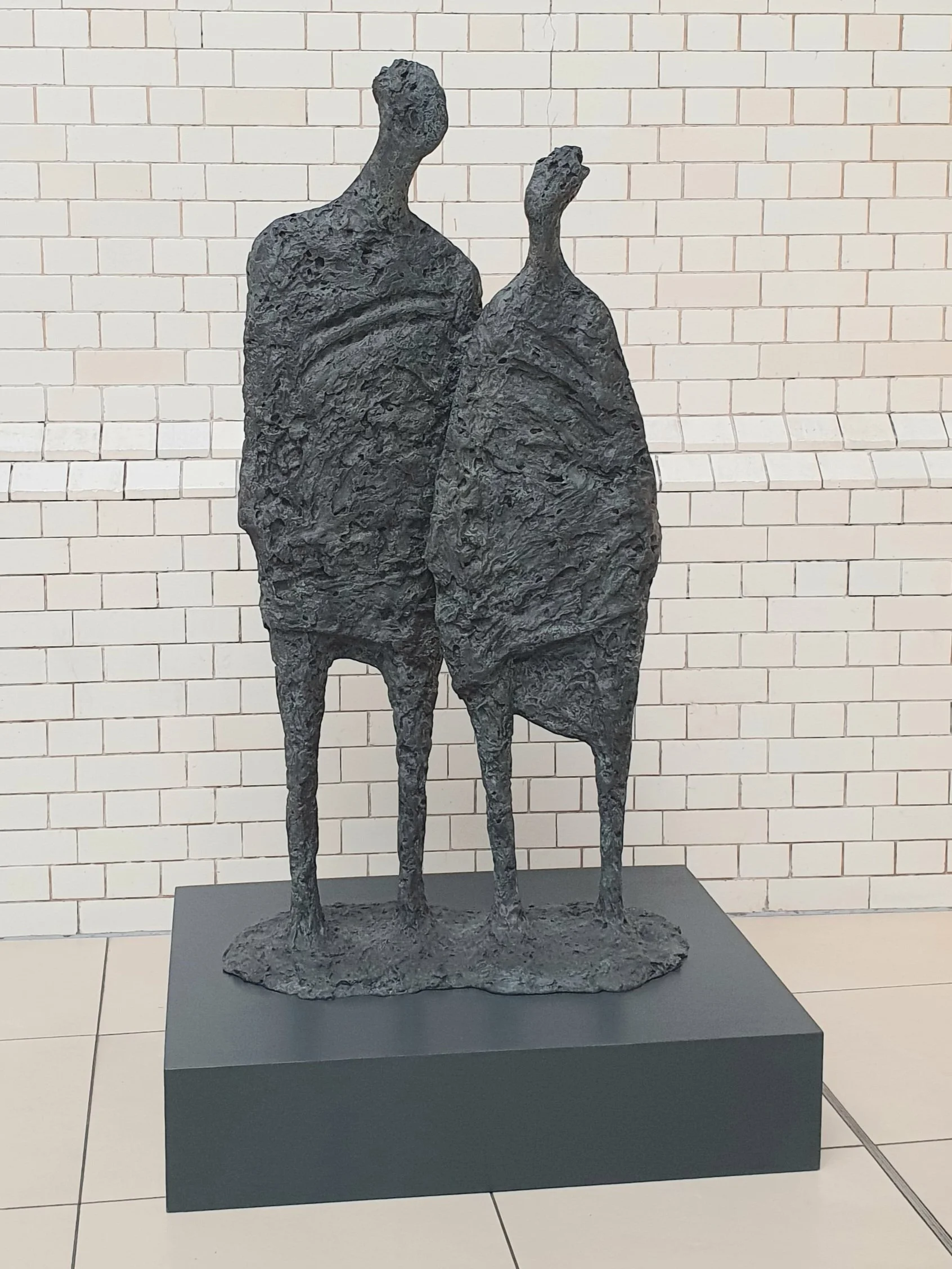 Metal sculpture of two elongated, abstracted figures, one taller and one shorter, standing side by side against a white brick wall.