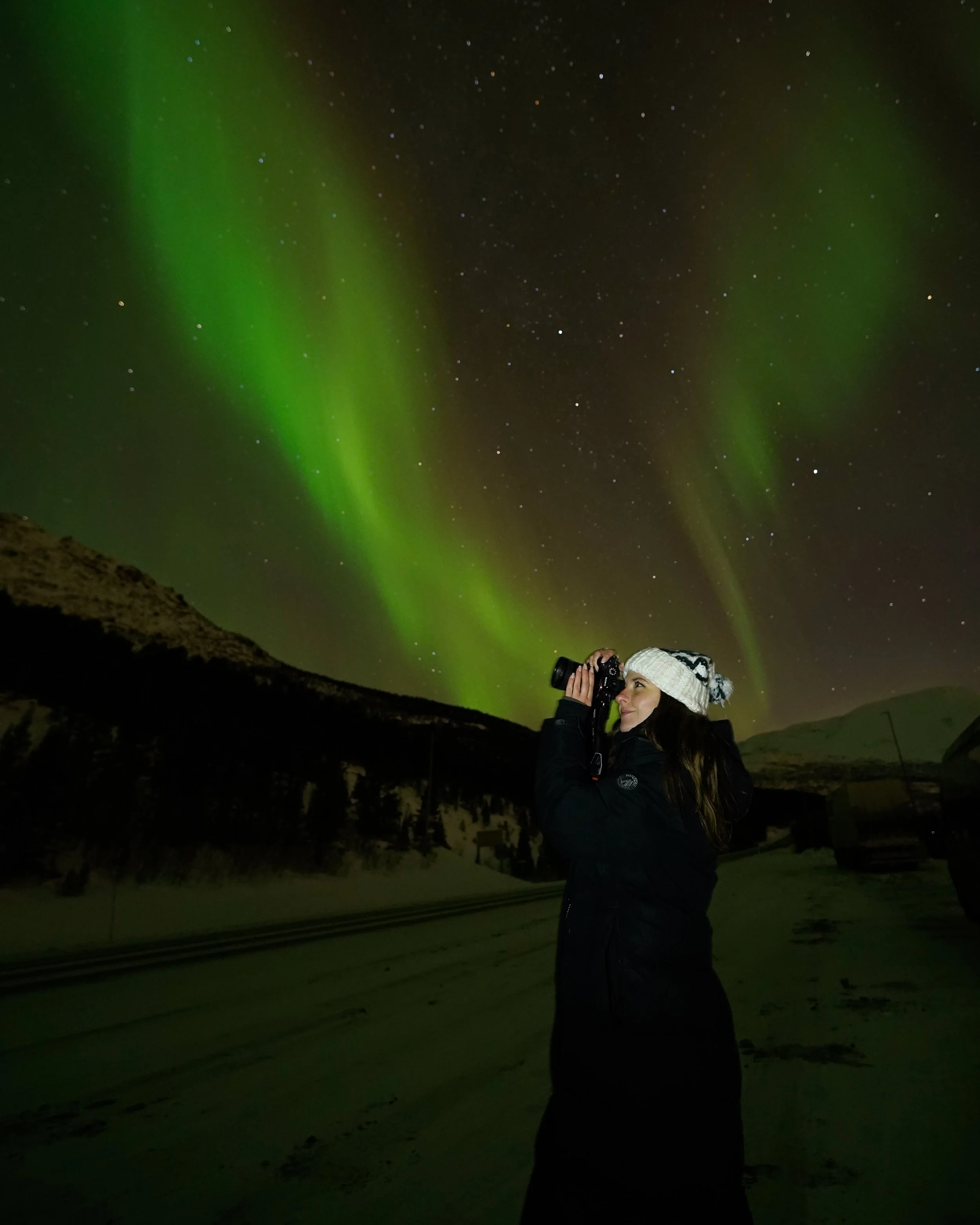 A woman holds a camera with the Northern Lights behind her
