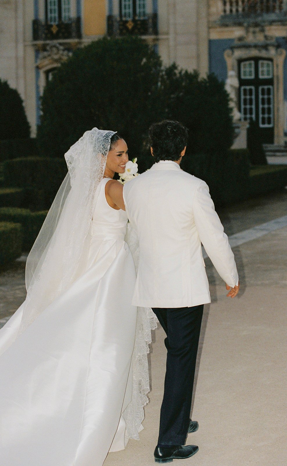 Newlyweds walking outside Palácio de Queluz, bride looking back smiling with white bouquet, analog film photo