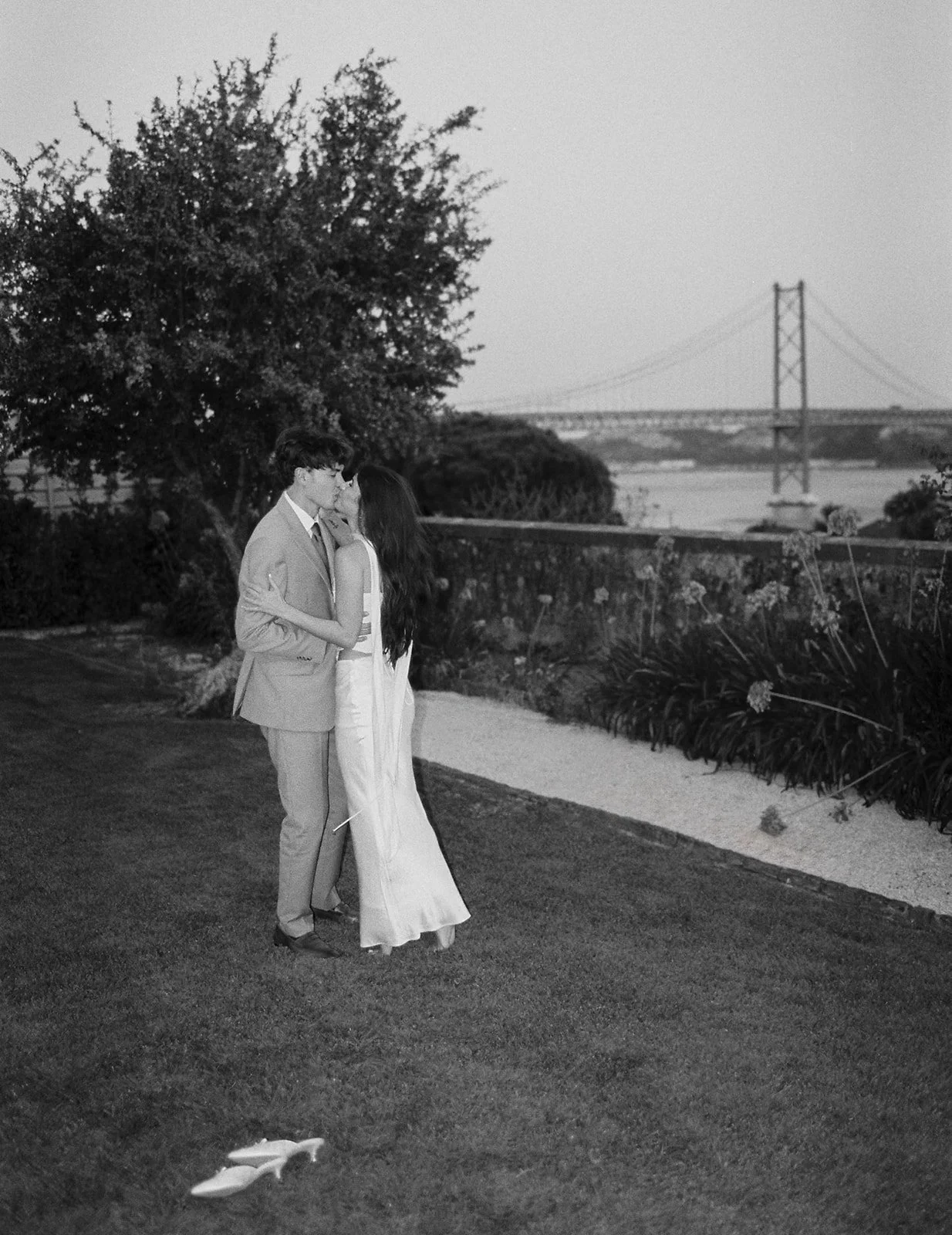 Couple sharing a quiet kiss in the garden of Pestana Palace Lisbon, with the 25 de Abril Bridge and the Tagus River visible in the background