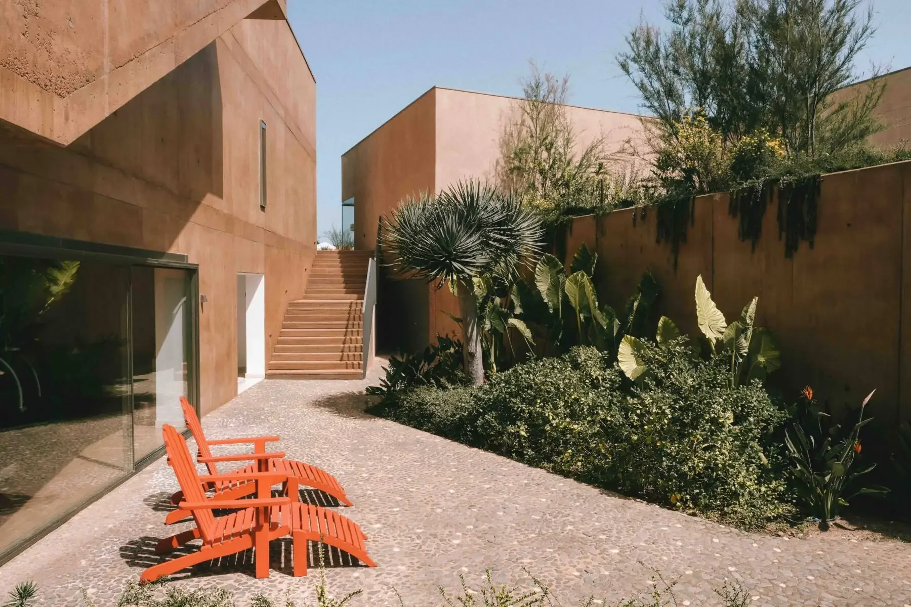 Modern courtyard with terracotta walls, lush greenery and orange lounge chairs at Praia do Canal Nature Retreat in Aljezur, Algarve.