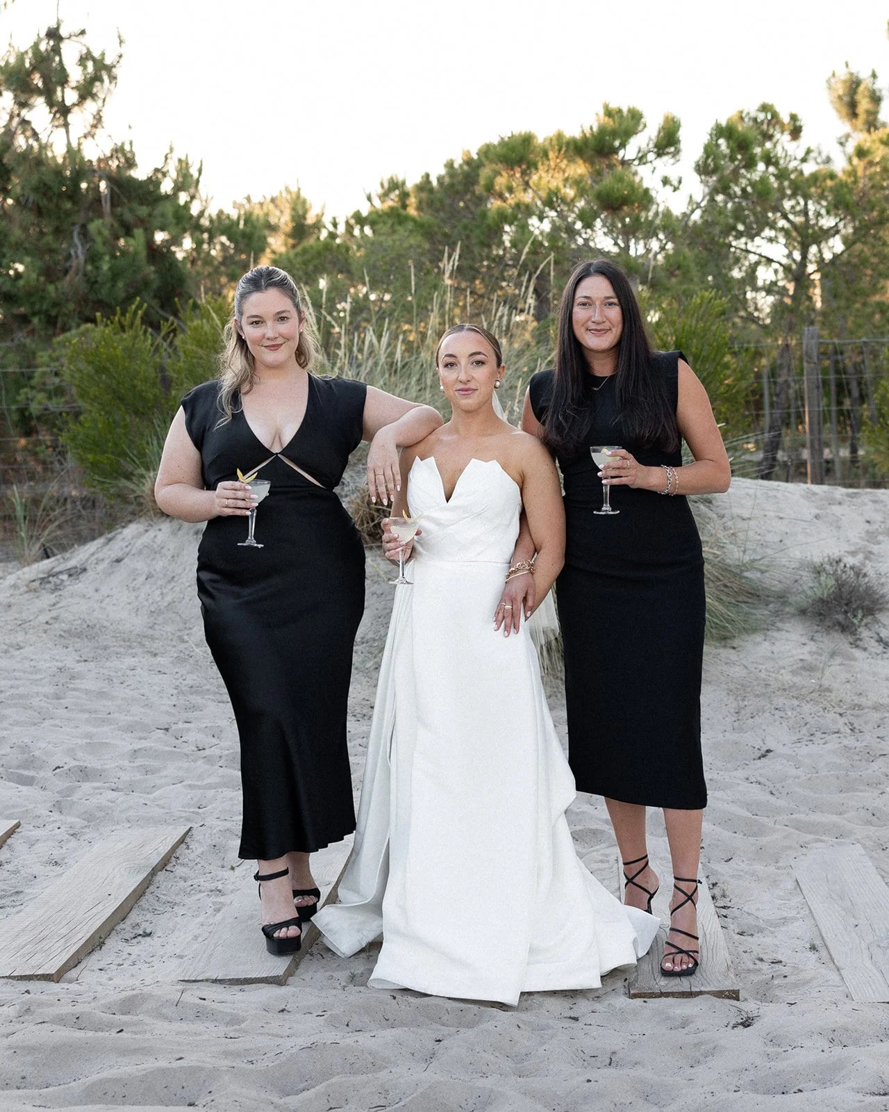 Bride and bridesmaids on a wooden boardwalk over sand dunes at golden hour, pine trees in background | The Lopes Photography