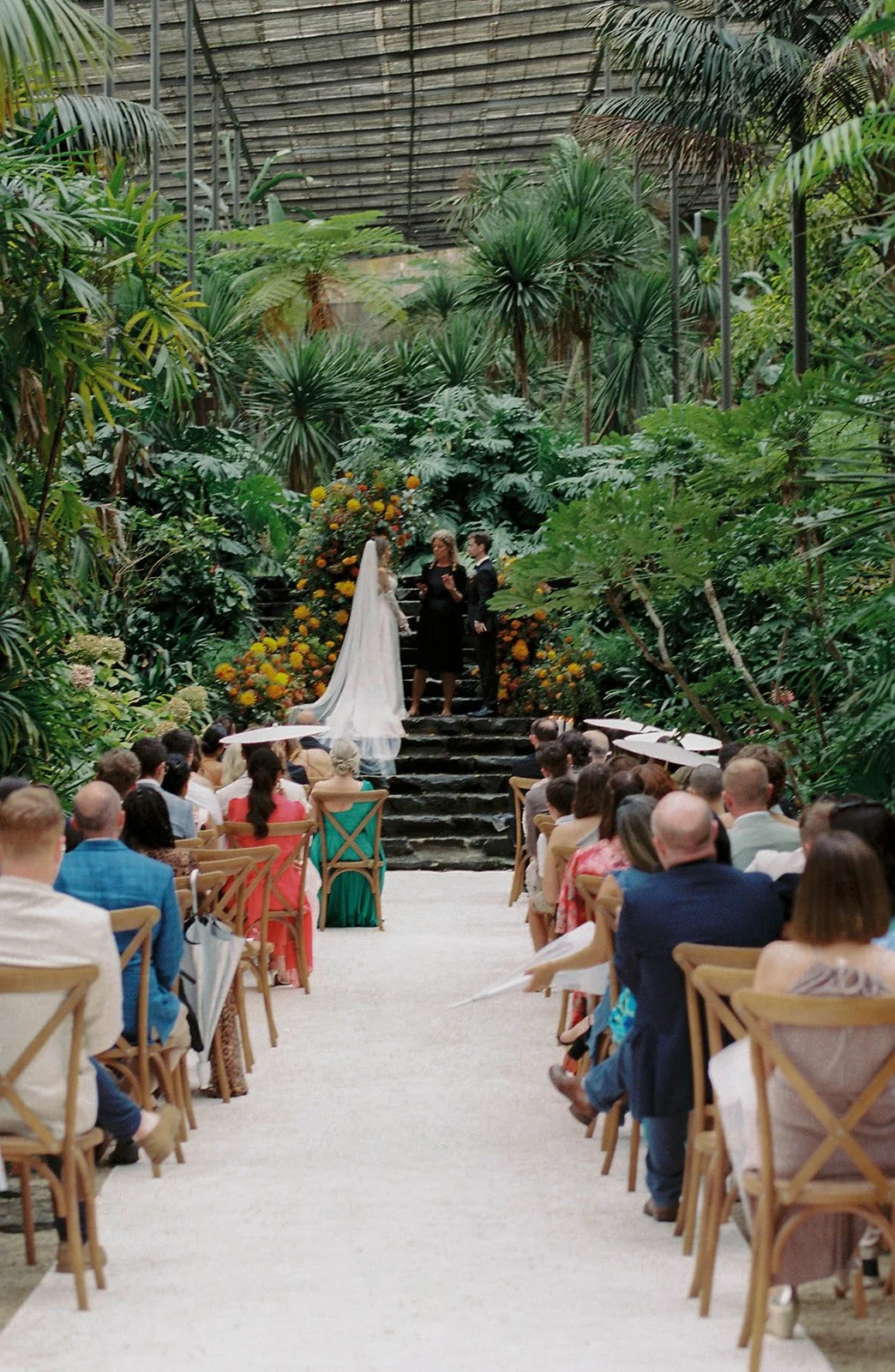 Wedding ceremony inside Estufa Fria in Lisbon, couple at the altar on stone steps surrounded by lush tropical vegetation with guests seated under white parasols along the aisle