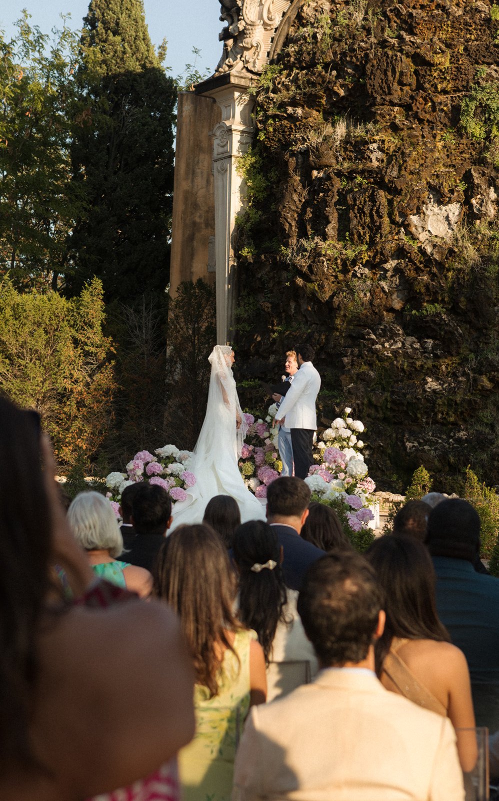 Wedding ceremony in the gardens of Palácio Nacional de Queluz, guests seated facing the historic stone monument in golden late-afternoon light, Portugal | The Lopes Photography