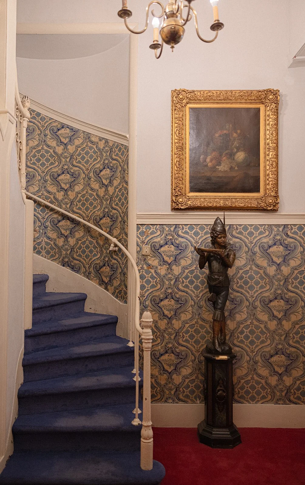 Grand winding staircase at Grémio Literário in Lisbon with blue carpet, ornate white iron banister, Portuguese azulejo tile walls, and a chandelier overhead
