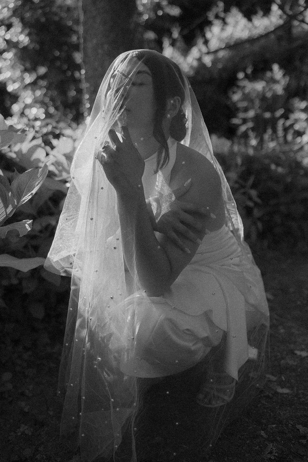 Film photo in black and white of a bride posing, close to the ground, with her veil on and eyes closed, while the sun is hitting her directly. Photo taken at Mosteiro de Landim in Braga.
