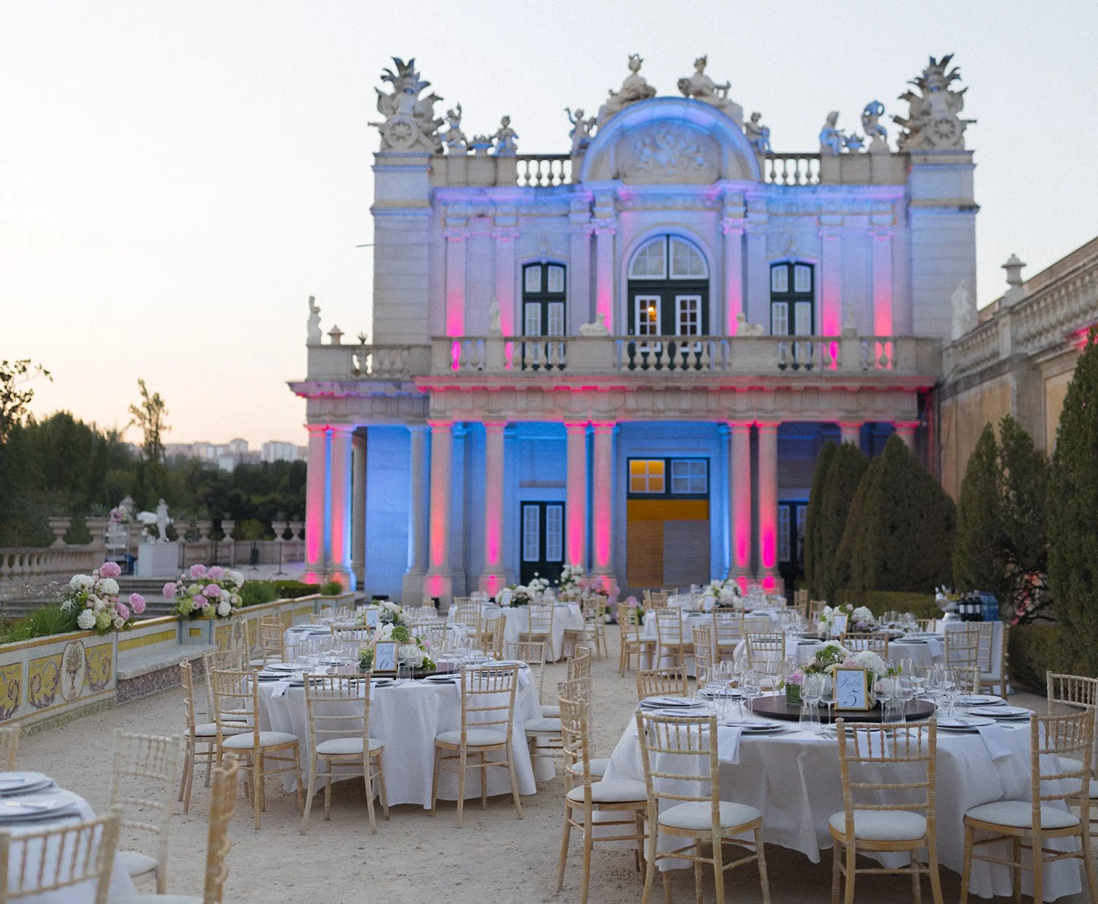 Outdoor wedding reception at a neoclassical palace in Portugal with tables arranged on an illuminated terrace at dusk._
