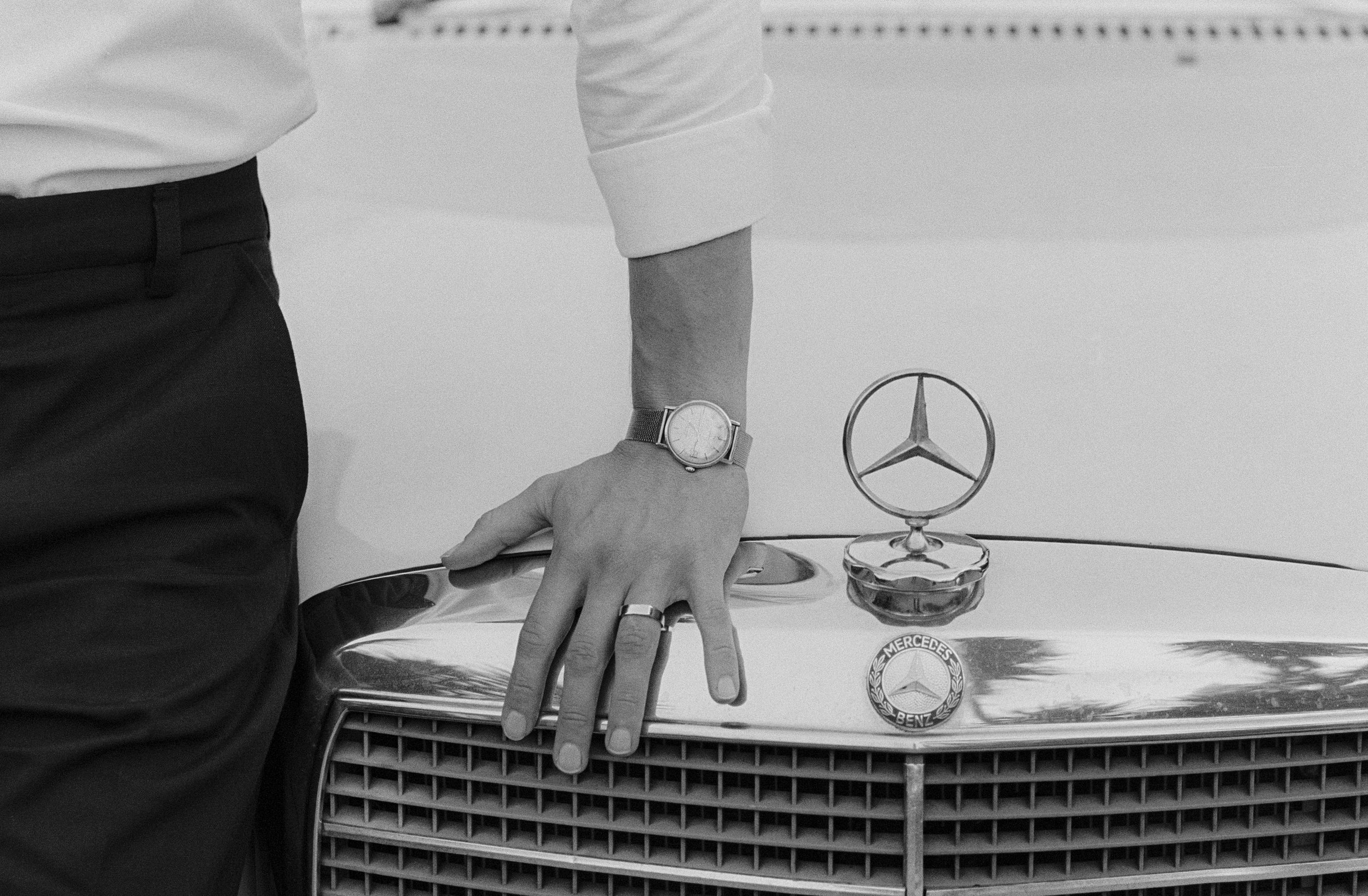 Black-and-white close-up of a groom’s hand resting on a vintage Mercedes grille and emblem.