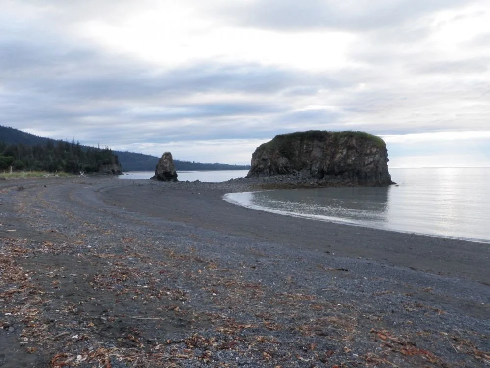 A wide-angle view of Haystack Rock in Kachemak Bay, Alaska. The large, flat-topped rock formation sits on a dark pebble beach at low tide, connected to the shoreline by a rocky tombolo. A smaller, jagged rock spire stands nearby. The scene features a