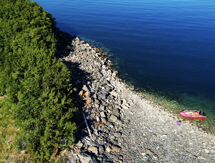 An aerial, high-angle view of Quarry Beach in Kachemak Bay, showing a rugged coastline where a dense, vibrant green forest meets a steep shore of light-colored rocks and grey boulders. The crystal-clear turquoise water reveals submerged rocks near th