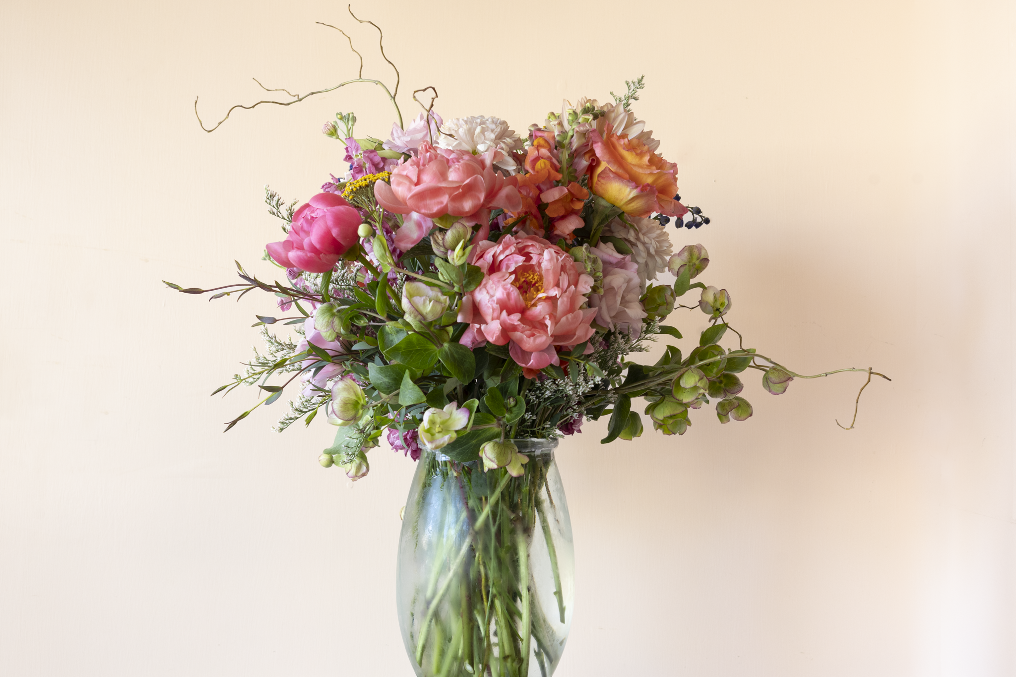 Designer's choice floral arrangement in tall, clear glass vase with a color palette of pinks, purples, creamy white, green, and oranges.