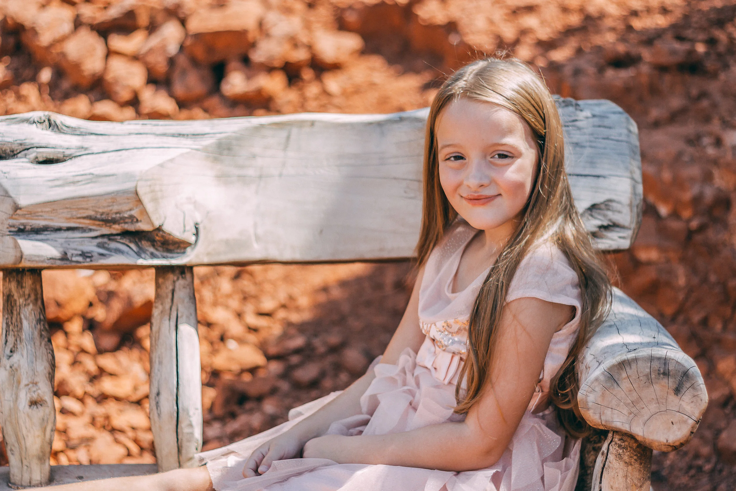 Portrait of a young girl seated on a rustic wooden bench against red rock, child photography by Everlux Photo Co.