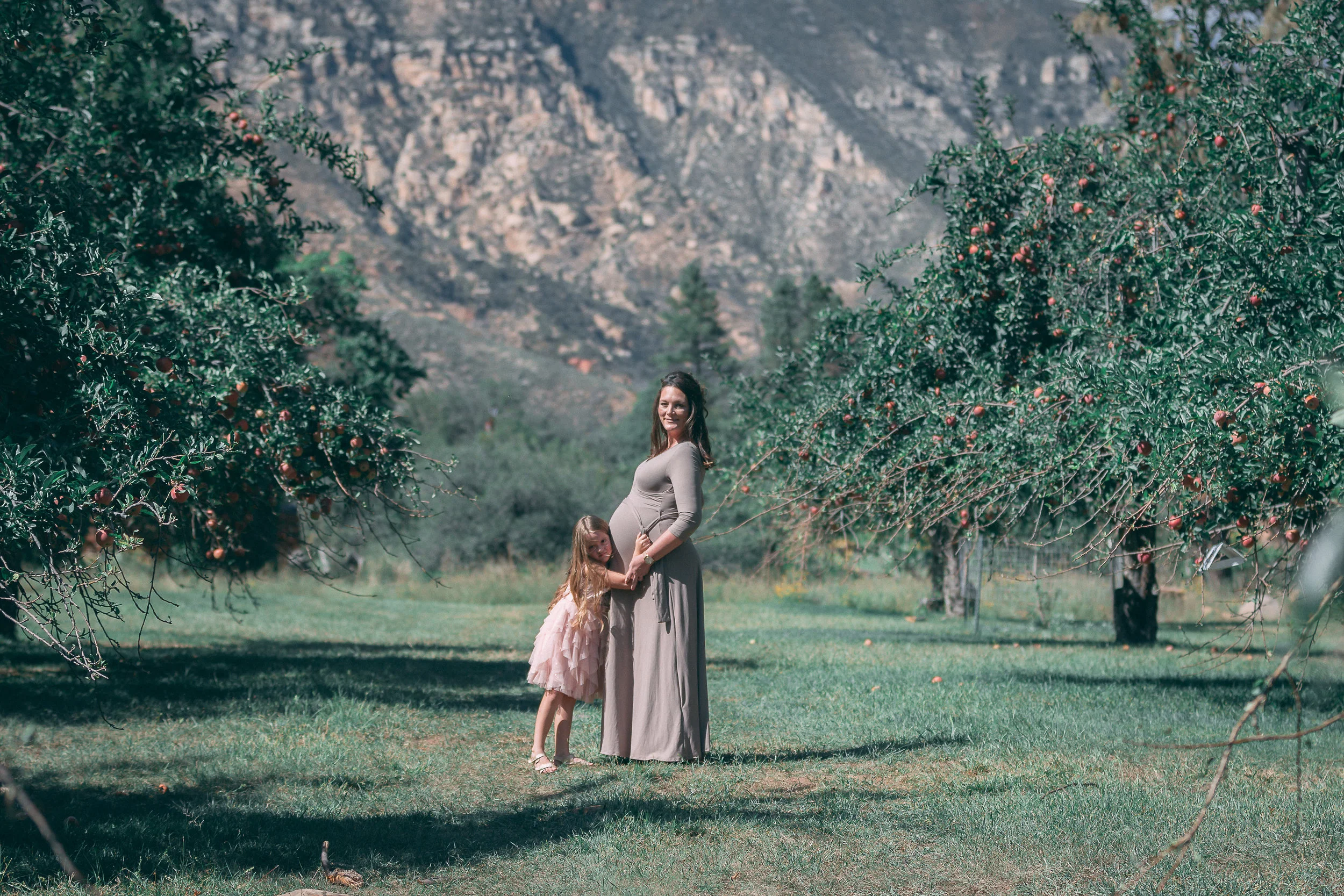 Wide maternity portrait of a pregnant woman with her daughter in a mountain meadow orchard, maternity photography by Everlux Photo Co.