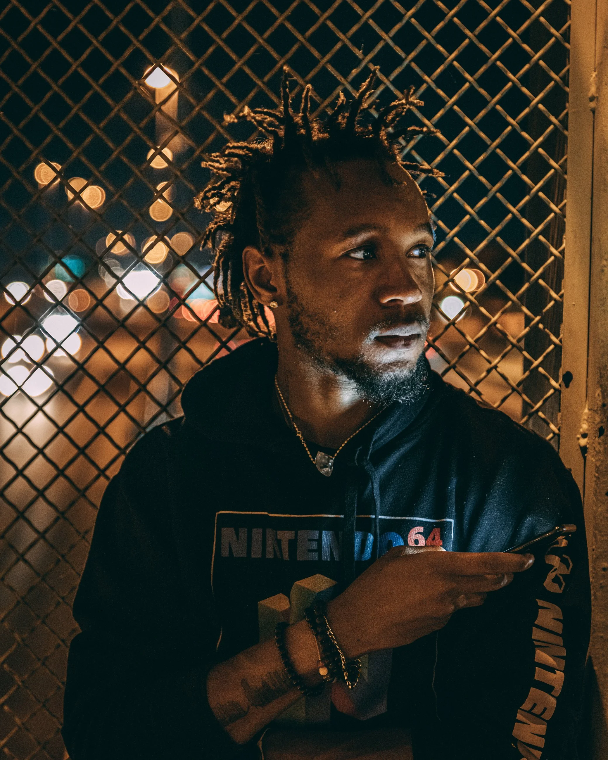 Nighttime portrait of a man by a chain-link fence with city lights behind, portrait photography by Everlux Photo Co.
