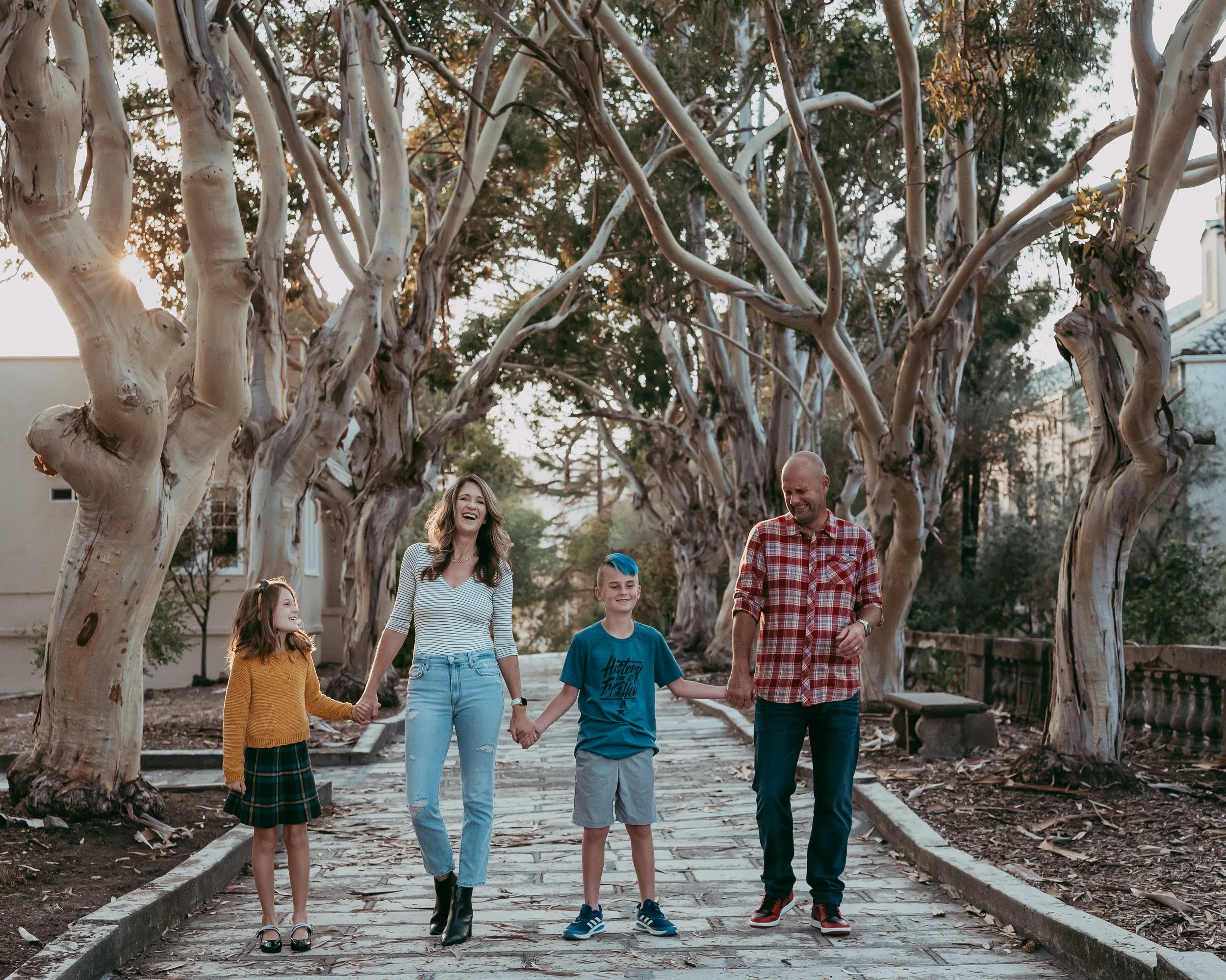 A happy family of four walking hand-in-hand down a tree-lined pathway in the late afternoon. The family includes a young girl, a woman, a young boy, and a man, all smiling and enjoying each other's company.