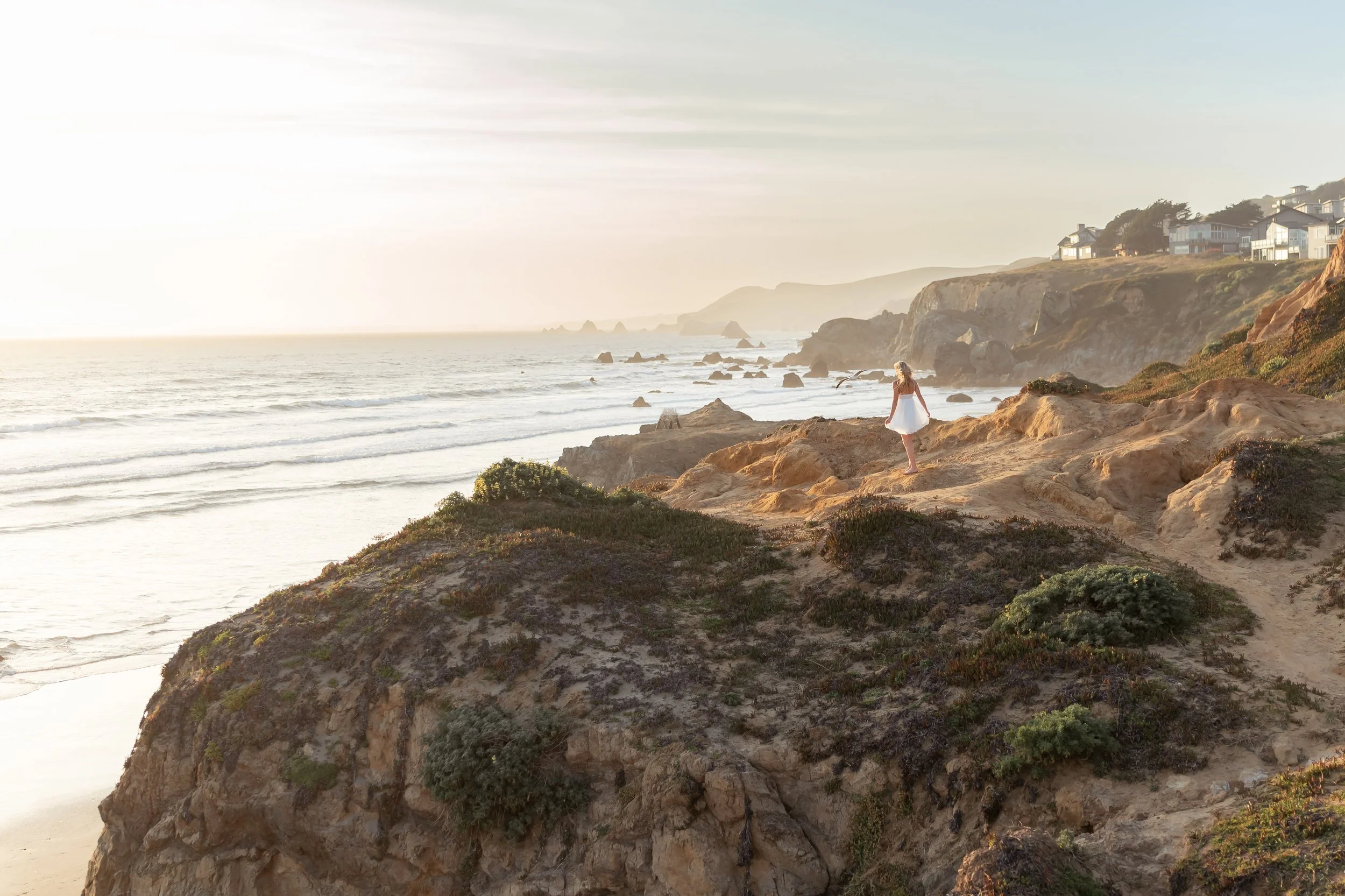 A woman in a white dress standing on a rocky cliff overlooking the ocean at sunset with houses on the hillside in the background.