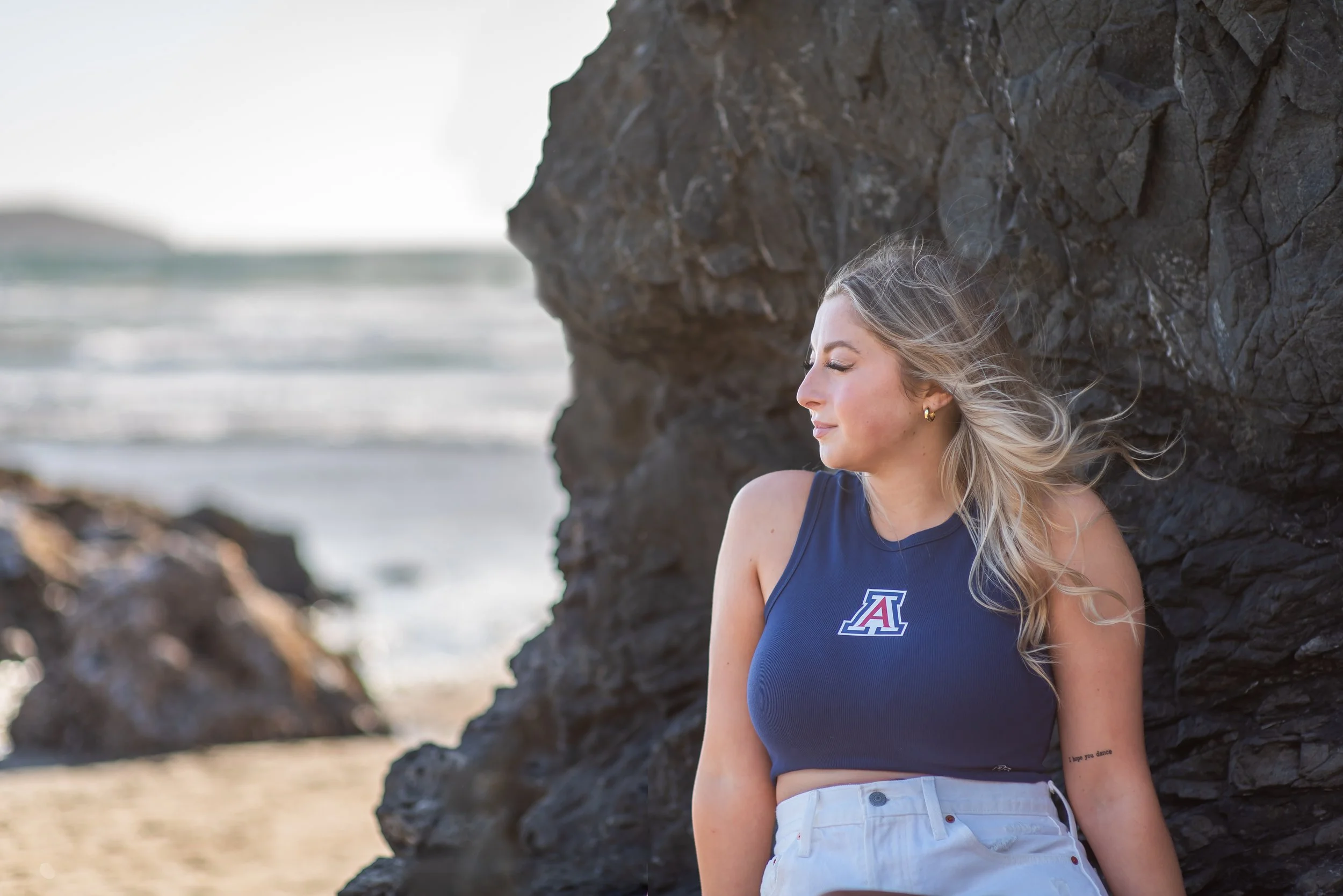 A young woman with blonde hair leaning against a large rocky formation on a beach, eyes closed, smiling, wearing a blue tank top with a University of Arizona logo, and white shorts, with a small tattoo on her arm reading 'I hope you dance'.