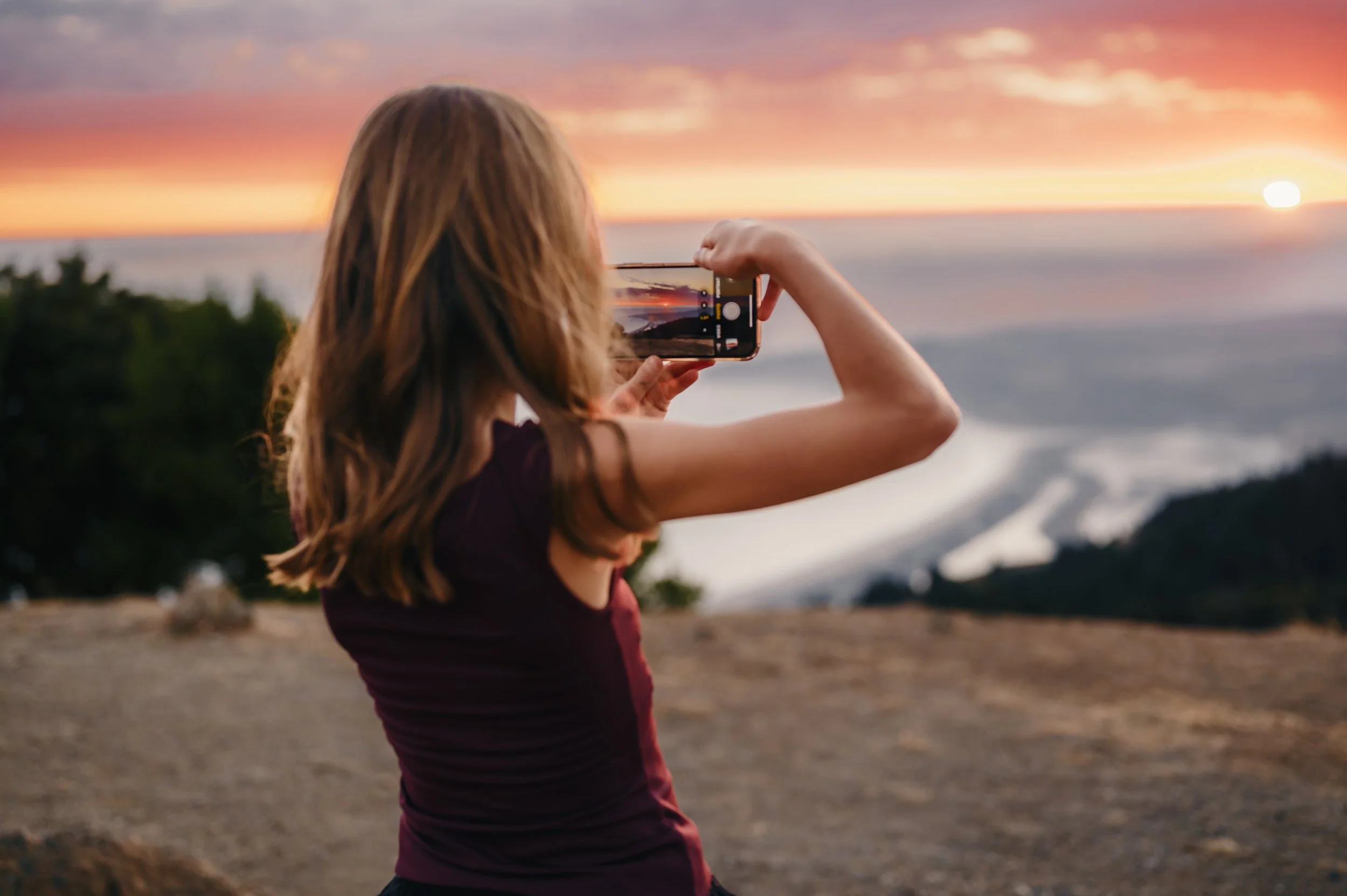 A woman with long hair holding a smartphone, taking a photo of a sunset over the ocean with a colorful sky.