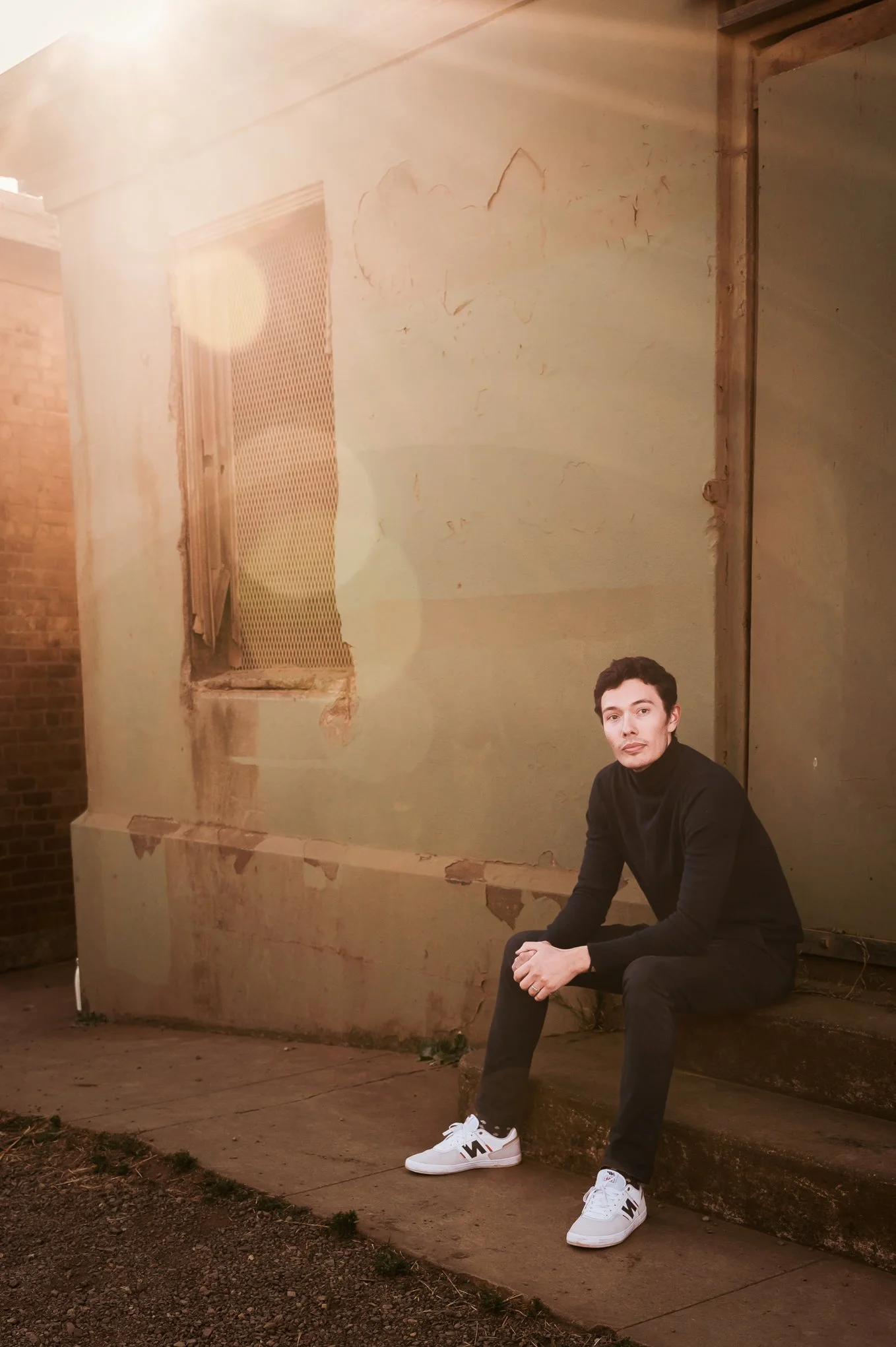 A young man in a black turtleneck and black pants sitting against a weathered wall with peeling paint and a window, illuminated by warm sunlight.