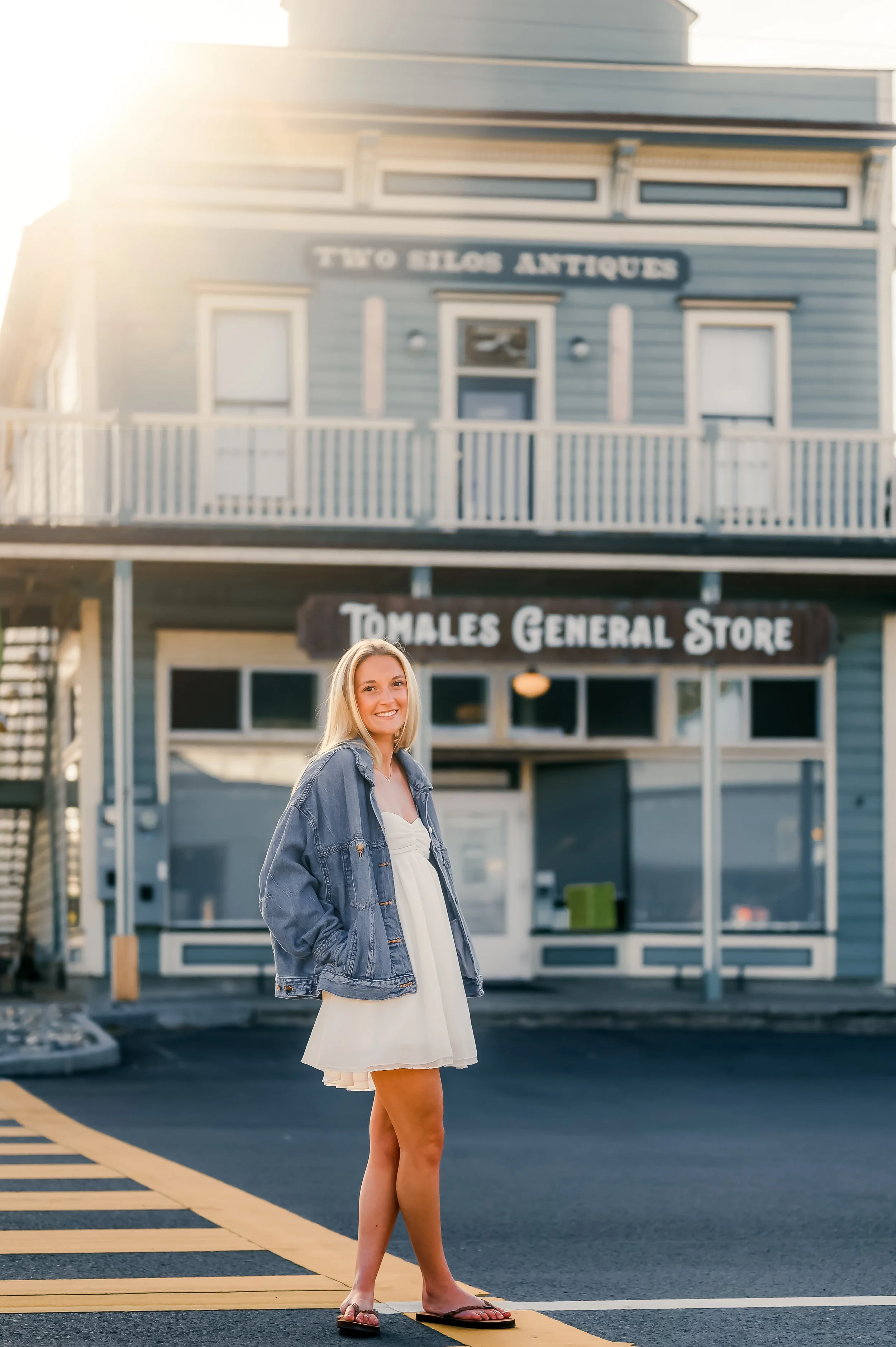 A young woman with blonde hair and a smile stands on a crosswalk, wearing a white dress, denim jacket, and flip-flops, in front of a two-story blue building with a sign that reads 'Two Silos Antiques' and another sign below that says 'Tomales General