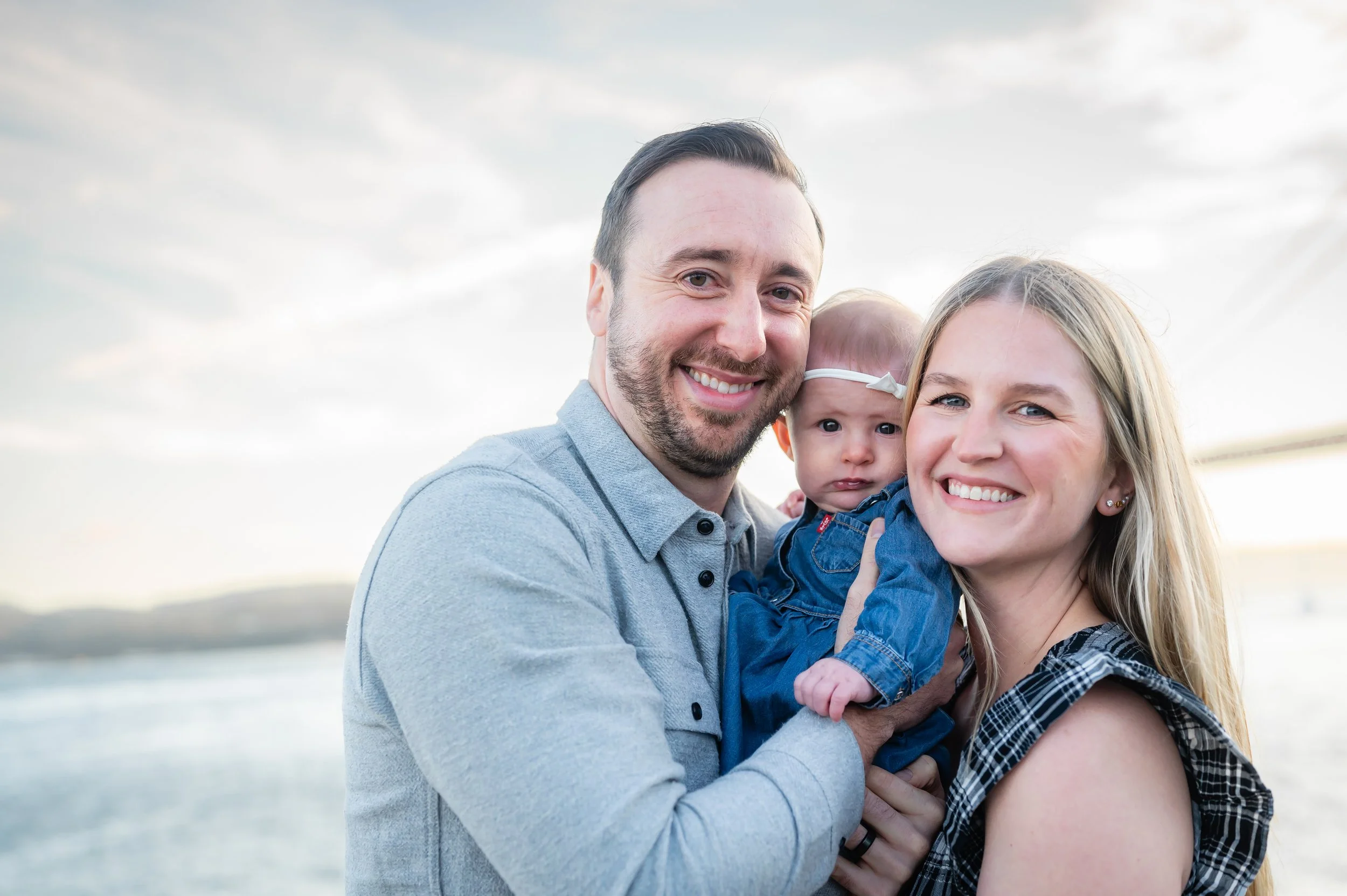 A happy family of three smiling by a body of water during sunset, with the father holding a baby girl and the mother standing beside them.