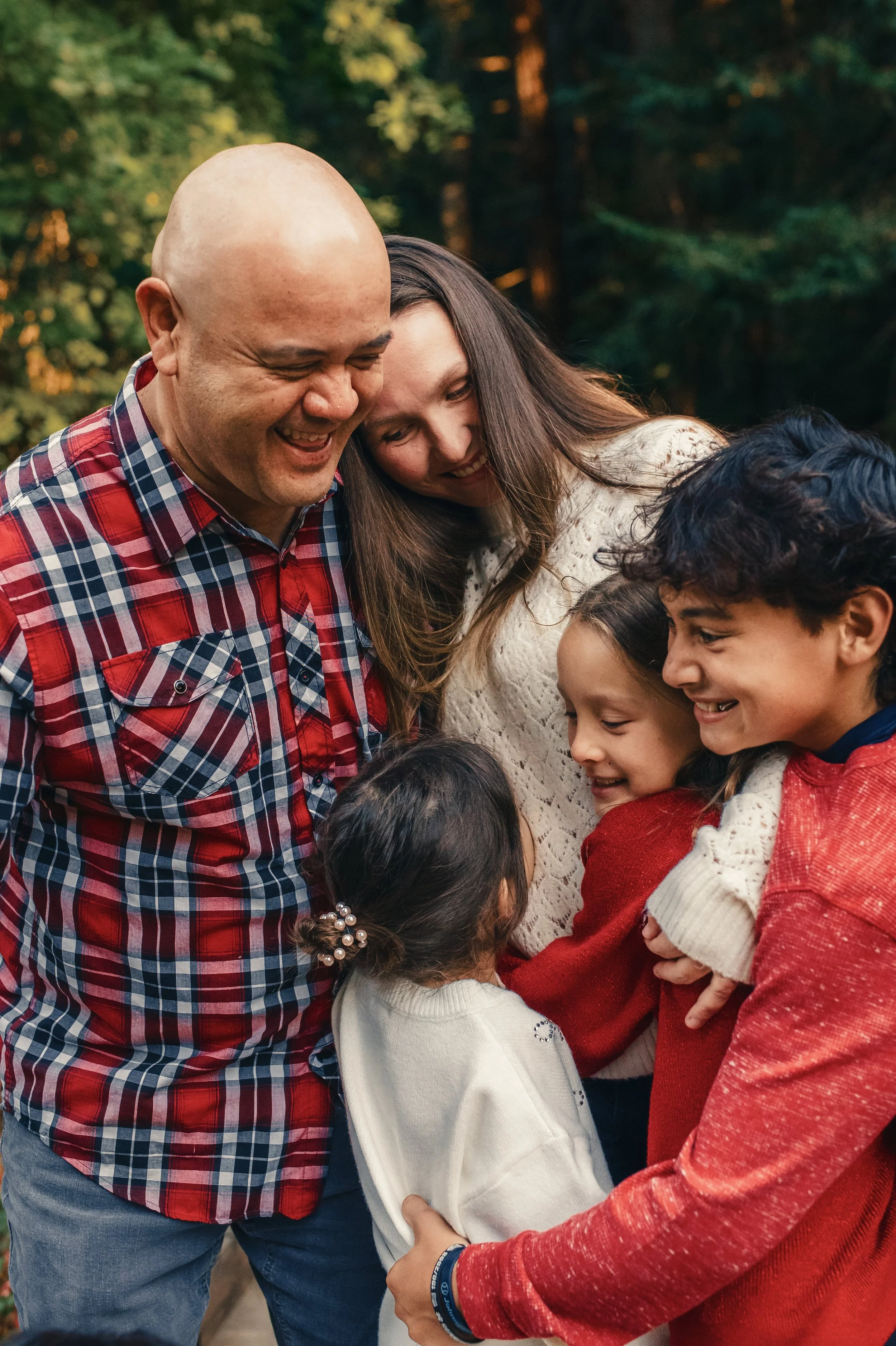 A group of people, including children and adults, are gathered closely together outdoors, smiling and embracing each other in a joyful moment.